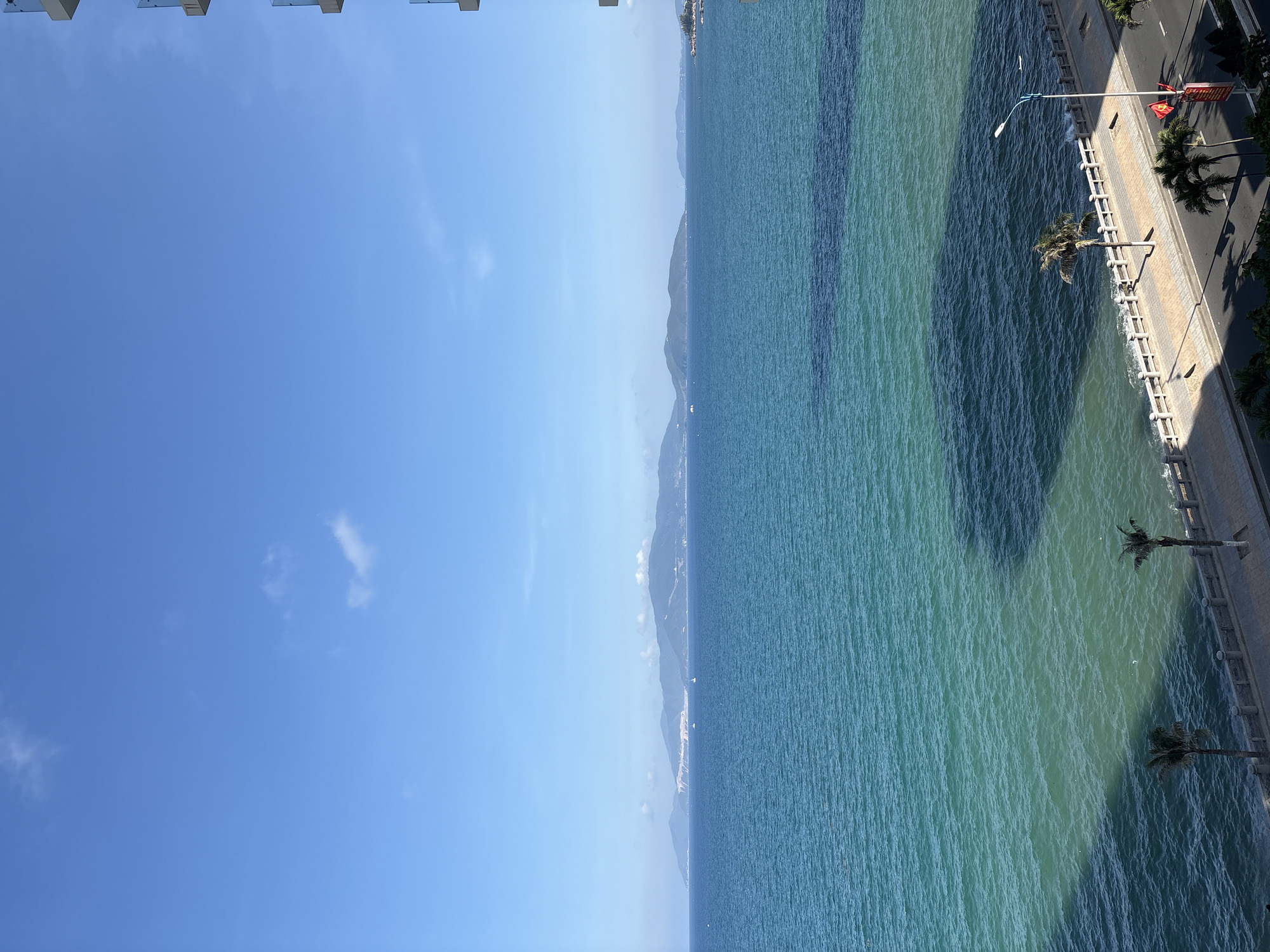This is an eye-level, outdoor shot of the coastline in Nha Trang, Vietnam on a sunny day. A blue sky with a few white clouds is on the left side of the image. The water is blue-green and extends across the right side of the image. Some coastal mountains are visible on the horizon. A narrow road lines the water with palm trees and streetlights along the edge. A few buildings are visible along the edge of the road.