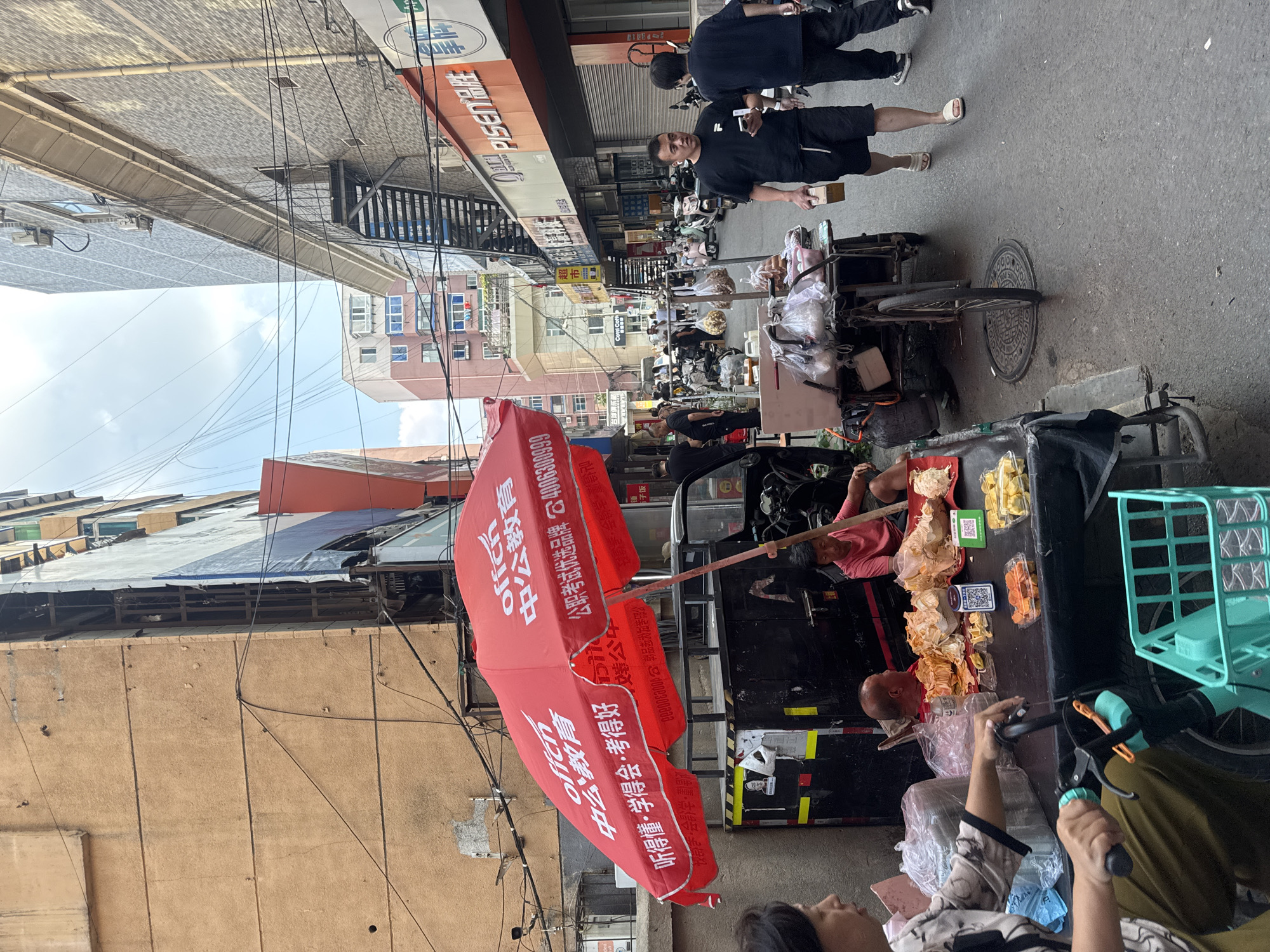The image depicts a bustling street scene in Dadong District, China. In the foreground, a woman on the left is seated behind a cart laden with various food items, including what appears to be seafood, dried fruits, and packaged goods. A red umbrella with Chinese lettering, advertising an educational institution, is prominently displayed over the cart. A man in a pink shirt is sitting on the cart behind the food. A turquoise bicycle is parked beside the woman. In the middle ground, a mobile food cart with three wheels is being pushed by an unseen person. Various people are walking along the street, some carrying bags or boxes. The street is lined with multi-story buildings, some appearing to be residential or commercial. Storefronts are visible, including one with an orange facade and Chinese signage. The street is paved with asphalt and there are power lines and air conditioning units visible on the buildings. A QR code is displayed on a small green item on the food cart, likely for payment. Two men in black t-shirts are walking away from the viewer towards the right side of the frame. The overall impression is one of everyday urban life and commerce in China.