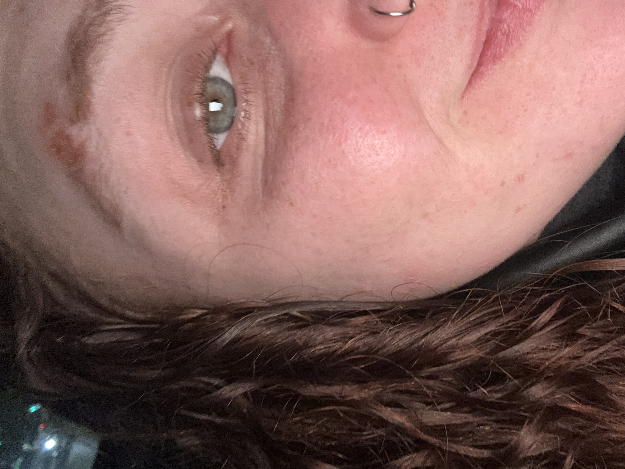 A close-up, angled image of a person's face, cropped from the forehead to the lower lip. The person has fair skin with freckles and a mole near their eye. Their eye is visible, with green iris and visible eyelashes. A silver nose ring is present. The person's hair is dark brown and appears to be damp. The background is dark and indistinct, with some blurred white and green lights visible in the bottom left corner. The image does not provide any information to indicate location in Buenos Aires, Argentina.