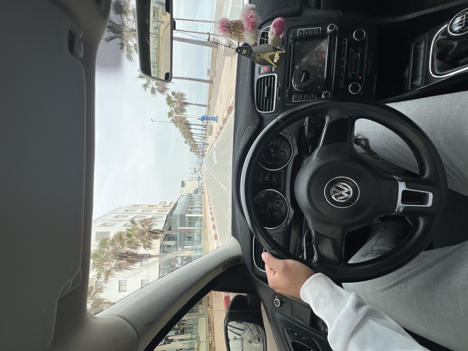The image offers a first-person perspective from the driver's seat of a car, looking out over a coastal road under an overcast sky. The driver's right hand, wearing a white long-sleeved top, rests on a black Volkswagen steering wheel, clearly showing the VW logo. The car's instrument panel indicates the time as 19:15 and the temperature as 25°C. The dashboard is visible, featuring a black cover and an infotainment system, along with decorative items such as a bird-like figurine and a pink fluffy pom-pom.
Outside the vehicle, a paved road extends into the distance, flanked by a red and white striped curb on the right, separating it from a wide, tiled promenade. Beyond the promenade, the sea is visible under the pale sky. The left side of the road is lined with numerous tall palm trees. Behind these trees, multi-story buildings, some with modern glass facades, stretch along the street. A sign on one of these buildings partially reads "Boulangerie" through its glass. A person dressed in blue workwear can be seen walking on the promenade in the mid-ground. The scene depicts a drive along a tranquil coastal area, consistent with the provided location context of Martil, Morocco, during a warm evening.
