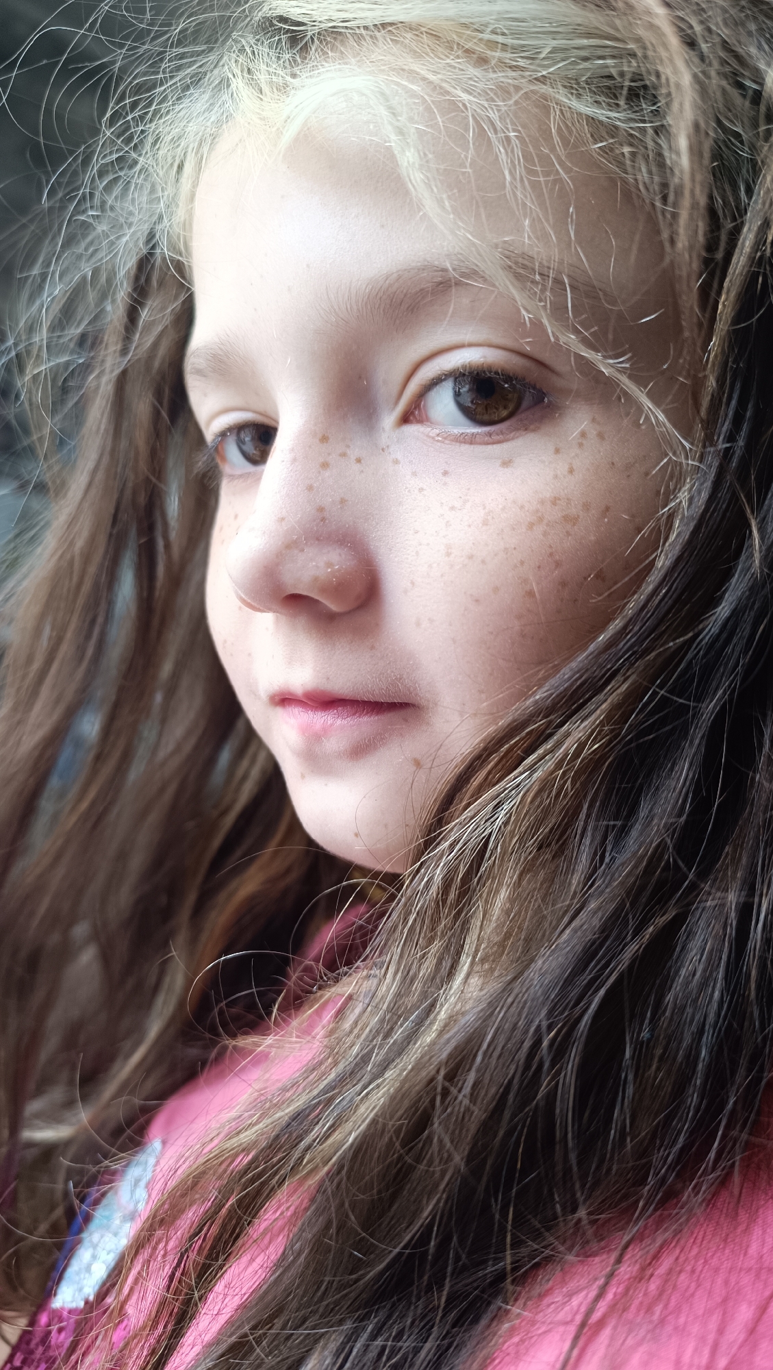 A close-up, profile shot of a young girl's face, with her head turned to the left. Her skin is fair with a scattering of light brown freckles across her nose and cheeks. She has large, brown eyes and full, pink lips. Her hair is long and wavy, with strands of both dark brown and blonde visible. She is wearing a pink top, part of which is visible in the lower right corner of the frame. The background is out of focus and appears to be a dark, possibly indoor setting. There is no information to indicate this image is from Karsha, Russia.