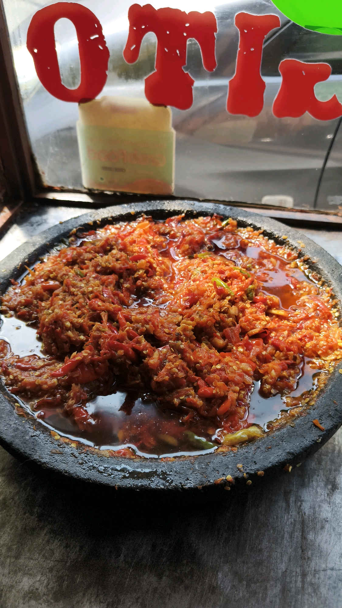 A close-up, overhead shot shows a mortar filled with a vibrant red chili paste, likely sambal, sitting on a weathered wooden surface. The paste is coarse and oily, with visible chili seeds and some green flecks of herbs or chilies. The mortar itself is made of dark, textured stone. In the blurred background, red lettering on glass appears to spell out "OTIK," suggesting a potential shop or eatery. A faded green label or advertisement is also visible behind the lettering. The lighting suggests an outdoor setting, possibly a street vendor's stall in Bekasi, Indonesia, given the context provided. No people are visible in the frame.