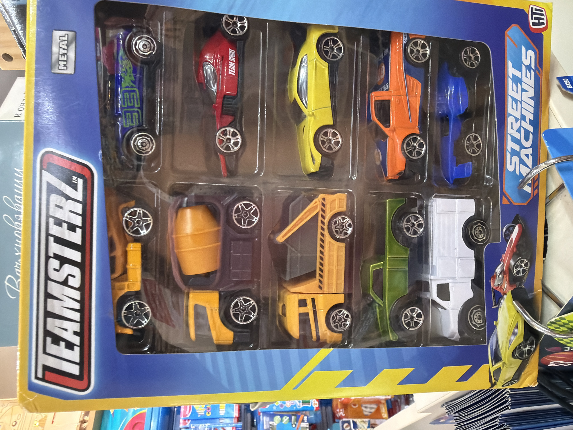 The image shows a package of Teamsterz Street Machines die-cast metal toy cars. The package contains ten different vehicles, including a blue sports car, a red race car with "TEAM SPIRIT" printed on the side, a yellow sports car, an orange van, a blue compact car, a yellow construction mixer, an orange crane, a green truck, and a white truck. The package is blue and yellow and has the Teamsterz logo in the lower left corner and the Street Machines logo in the upper right corner. The package appears to be located on a shelf in a store. While the description requested location information in Plovdiv, Bulgaria, there is no evidence to infer this from the image.