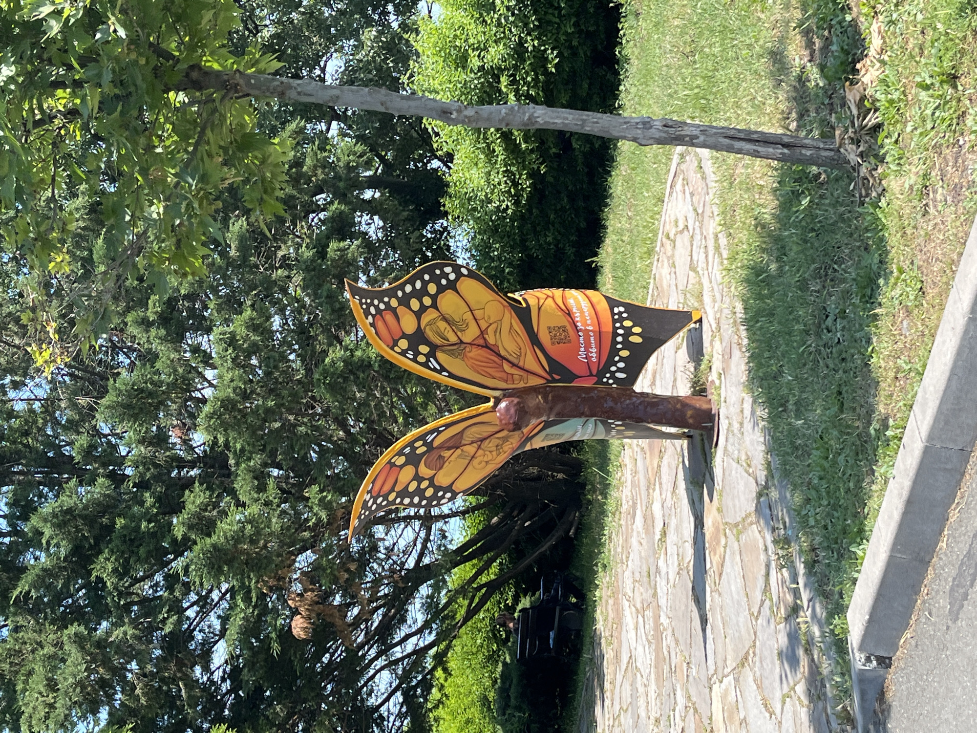 A large, colorful butterfly-shaped sculpture, likely functioning as a bench, is prominently displayed outdoors on a sunny day. The sculpture is painted predominantly in shades of orange, yellow, and black, resembling a Monarch butterfly. Its wings feature intricate artwork depicting a mother embracing a child, with additional figures and swirling patterns.
On the upper wing, Cyrillic text is visible, which reads: "Мама, татко, обичаме ви!" (Mom, Dad, we love you!) and beneath it "Минало, настояще и бъдеще" (Past, present, and future). A QR code is also present on the upper wing.
The butterfly is situated on a light-colored, irregularly paved stone path, possibly flagstone or cobblestone. To the right of the path is a strip of green grass, bordered by a concrete curb and an asphalt road. A variety of trees and dense green foliage form the backdrop, suggesting a park or public garden setting. The sun is bright, casting distinct shadows on the ground and indicating daytime. The scene conveys a peaceful, public recreational area, likely celebrating family or remembrance, given the text on the sculpture.