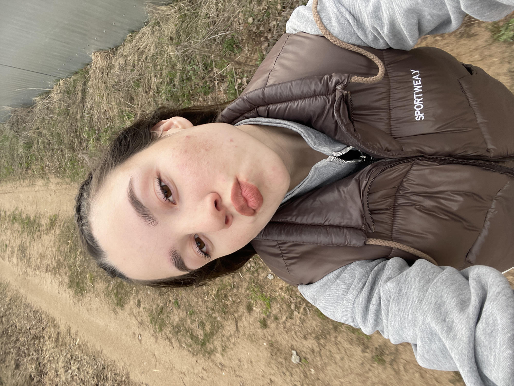 The image shows a close-up selfie of a young person, likely female, captured outdoors. The subject has dark hair, brown eyes, and prominent eyebrows, looking directly towards the camera with a pouting expression. Some skin texture and minor blemishes are visible on their face.
They are dressed in a brown, shiny puffer vest worn over a light grey hooded sweatshirt, the sleeves of which are bunched up. A brown braided drawstring is visible near the neck area of the sweatshirt. The vest features white text on the right side of the chest that clearly reads "SPORTWEAY".
The background suggests an outdoor, possibly rural or semi-rural setting. A dirt path or track runs across the lower and left portions of the image. Beyond this, there is dry, yellowish-brown grass and sparse vegetation, indicative of early spring or late autumn. In the upper left corner, a section of a grey, corrugated metal fence or wall can be seen. The lighting is natural and appears to be daylight.
The activity is a self-portrait or selfie. The location context for the image is Ольховка, Russia.