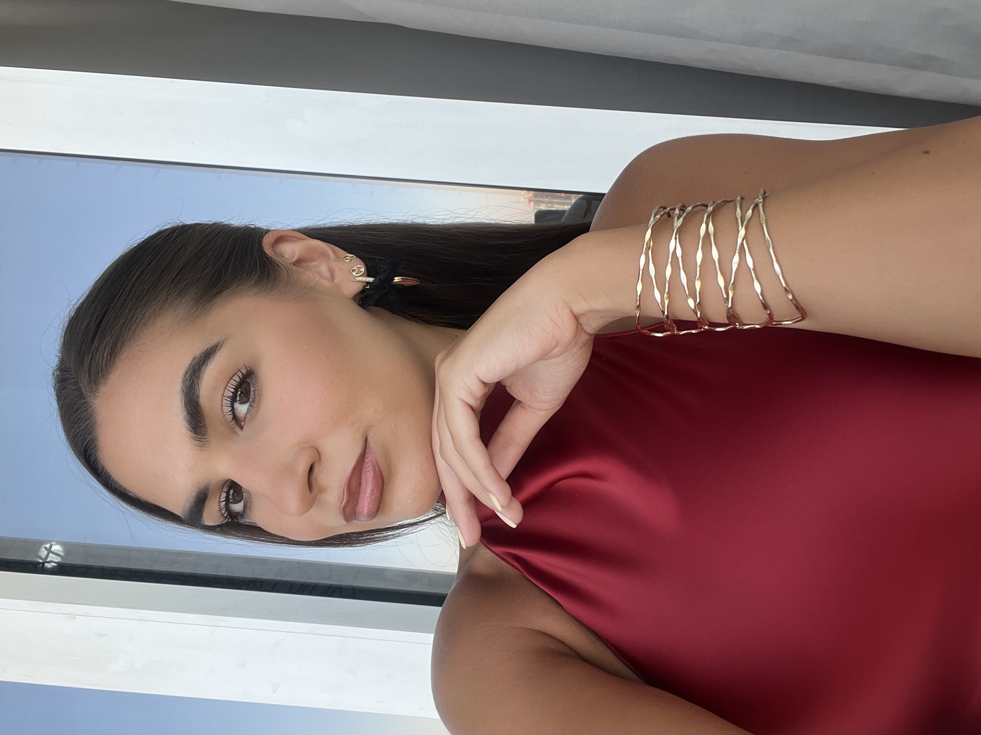 A fair-skinned woman with brown hair, wearing a red satin top, is posing indoors. She has on multiple gold bracelets and earrings. Behind her is a window with a view of what appears to be Dubai's skyline.