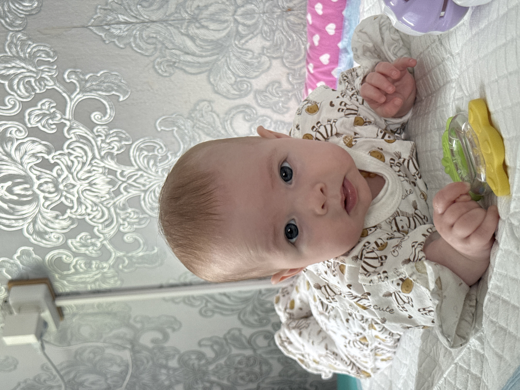 A baby wearing a zebra-patterned onesie is lying on a white quilted surface, possibly a blanket or playmat. The baby is holding a green and yellow rattle toy with their right hand. The baby's head is turned towards the viewer, and they have light-colored hair and blue eyes. The background consists of a wall with a floral pattern and an electrical outlet. A portion of a pink and white heart-patterned blanket is visible above the baby's head. The location is Кемуль, Russia.