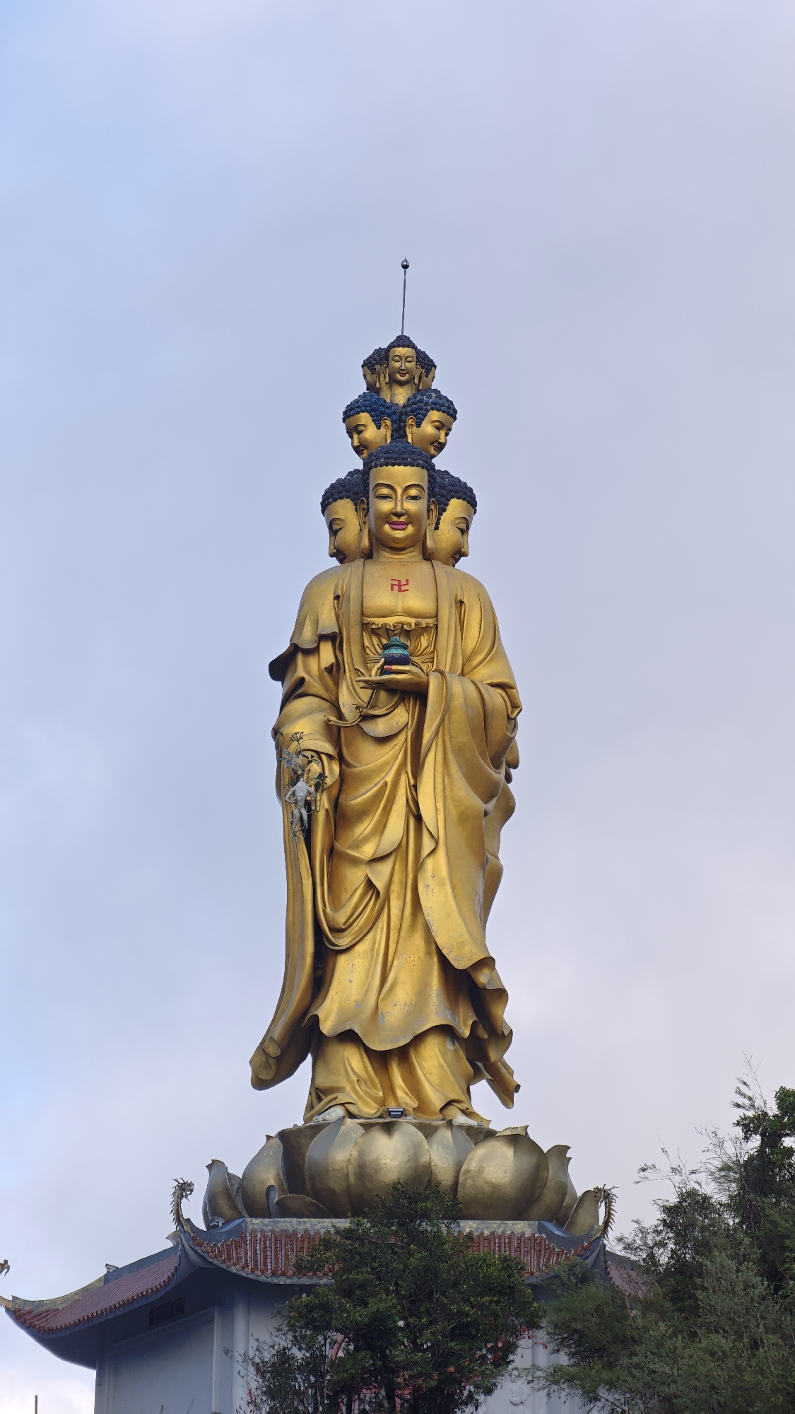 A golden statue of Buddha with multiple heads stands on a golden lotus pedestal. The statue is set against a cloudy sky. At the bottom of the lotus pedestal, the corner of a building is visible, with a roof adorned with dragon heads. Bushes and trees are seen on the right side of the frame. The statue is located in Binh Lu, Vietnam.