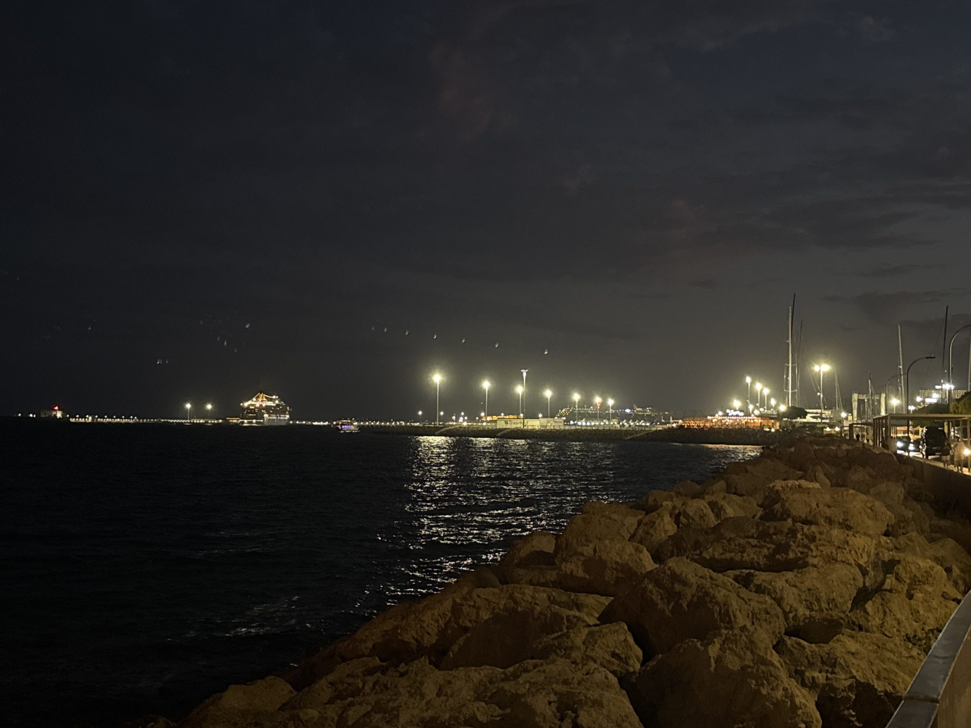 At night, a wide shot captures a harbor in Palma, Spain, from a rocky embankment. The dark, calm water reflects the illuminated city lights and a large cruise ship docked in the distance. The cruise ship is brightly lit with multiple decks visible. A smaller boat is also visible near the cruise ship. The background shows a long pier lined with streetlights, buildings, and docked sailboats. The sky is dark with some clouds. The foreground consists of large, rough-textured rocks of various sizes, with a metal railing visible on the far right. The overall scene conveys a sense of nighttime activity and illumination in a coastal city.