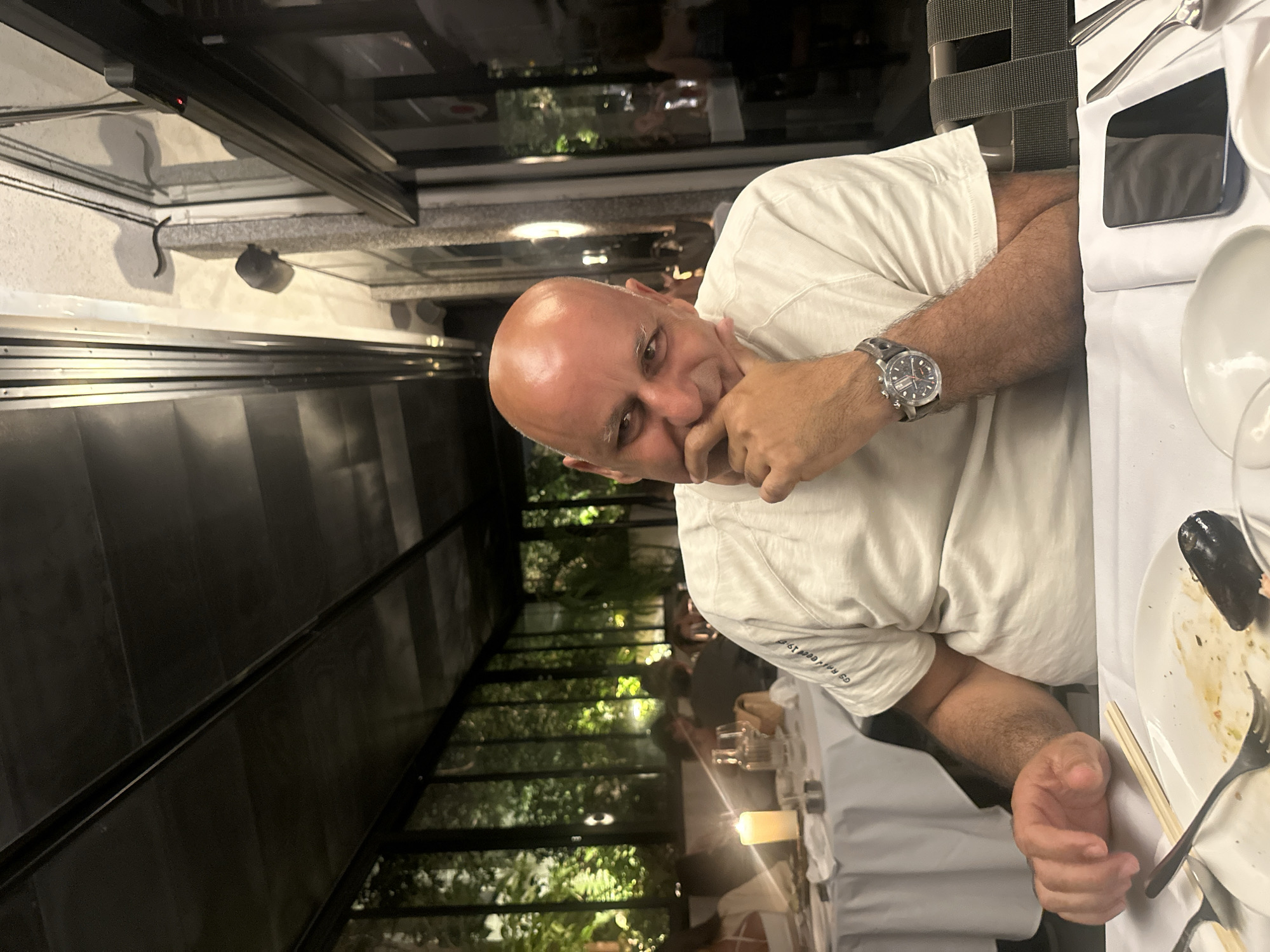 A bald man with fair skin sits at a table in an upscale restaurant, appearing to be in Tel-Aviv, Israel, judging by the decor and style. He wears a white t-shirt and a watch, and he has his hand raised near his face. The table is set with white linen, plates, cutlery, and a phone. A candle illuminates the scene. The restaurant has a modern aesthetic with a glass wall that overlooks green foliage. Other diners are visible in the background.