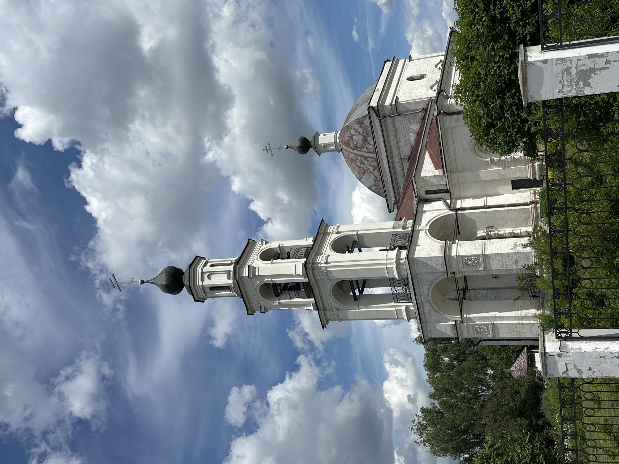 The image shows a white stone church with a tall bell tower and a dome topped with a cross, set against a blue sky with fluffy white clouds. The church has multiple arched windows and decorative architectural elements. In the foreground, there is a black metal fence and a white stone pillar. Lush green trees and bushes surround the church, with a small red-roofed structure visible behind the fence on the left. The overall scene depicts a serene religious building in a verdant setting. This church is likely located in the village of Ерино, Russia, as indicated by the prompt.