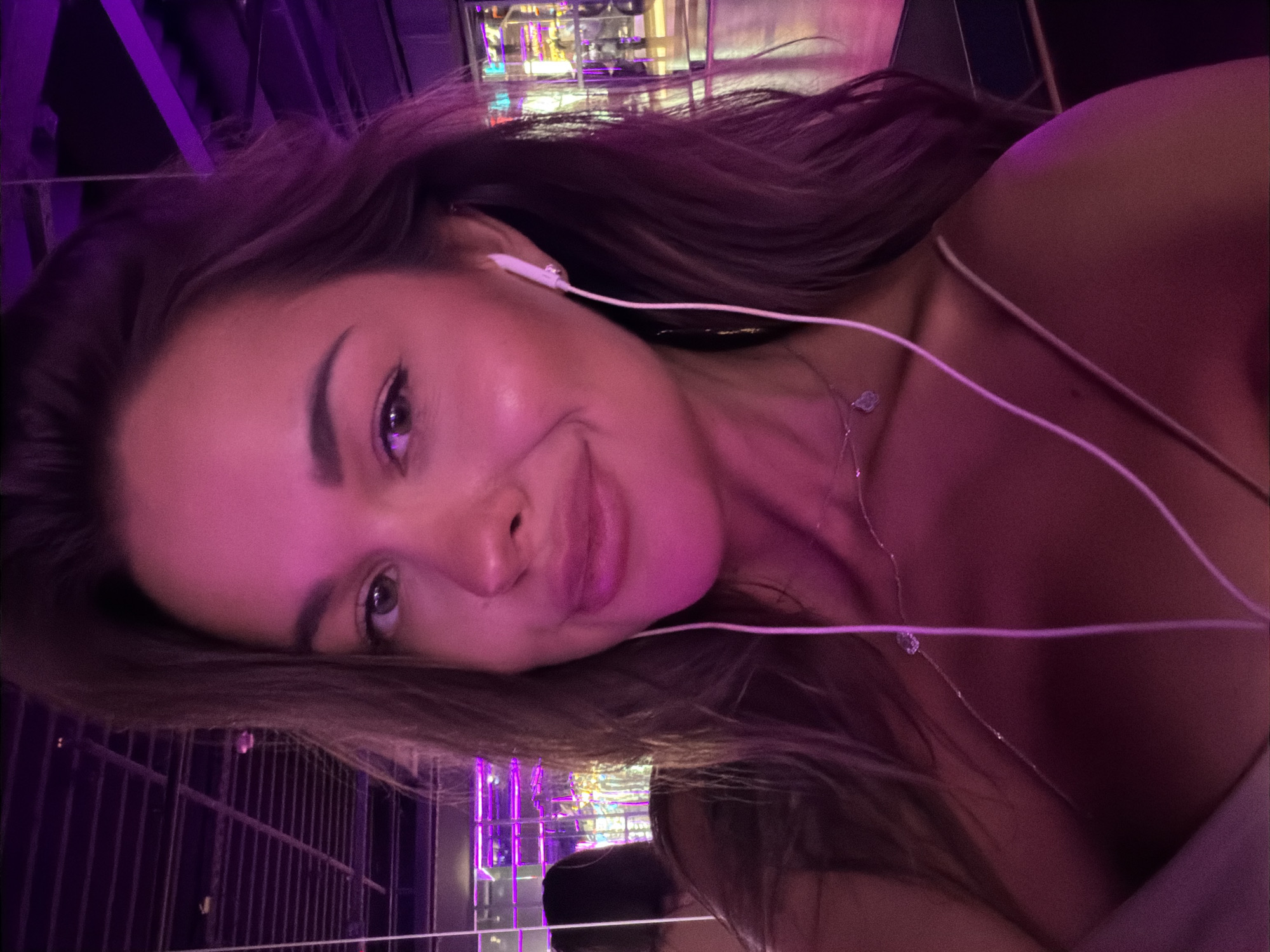 A young woman is taking a selfie with headphones in her ears. She is wearing a thin-strapped top and a delicate necklace. The background suggests an indoor setting with purple lighting and reflections, possibly a club or entertainment venue in Saint Petersburg, Russia.