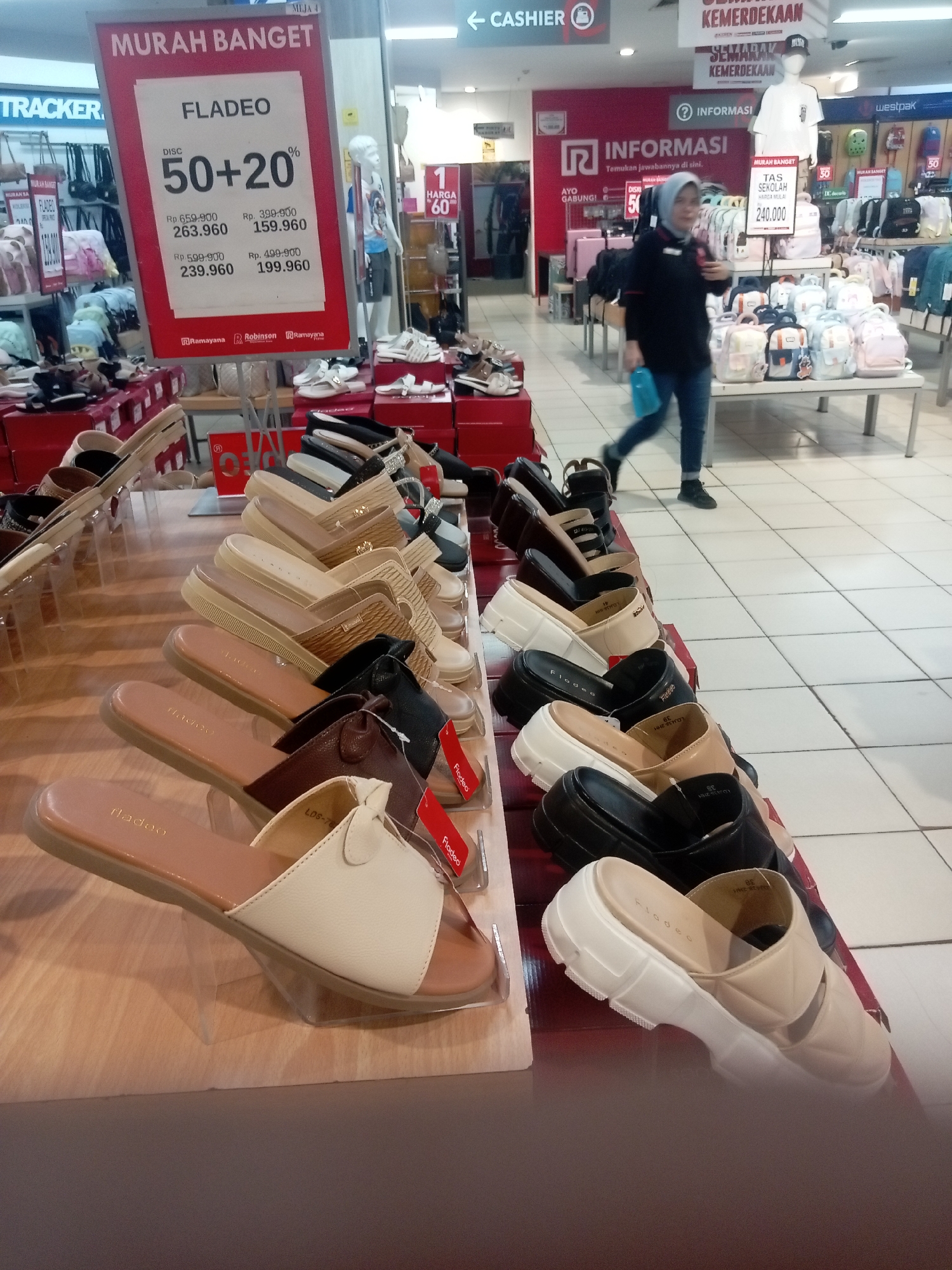 The image shows a retail display of women's footwear, likely in a department store, in Batam, Indonesia. The display features several rows of sandals and platform shoes on wooden shelves and clear acrylic stands. A large red sign prominently advertises a "MURAH BANGET" (very cheap) sale for the brand "FLADEO," offering discounts of "50+20%." Below the discount, specific prices are listed: Rp 263.960 (originally Rp 659.900), Rp 159.960 (originally Rp 399.900), Rp 239.960 (originally Rp 599.900), and Rp 199.960 (originally Rp 499.900). The background shows more merchandise, including bags, and a blurry figure of a woman in a hijab walking away from the camera. Signage indicating "CASHIER" and "INFORMASI" (information) is visible in the upper and mid-ground, suggesting a typical mall or department store environment. The floor is tiled with a white and light grey checkered pattern.