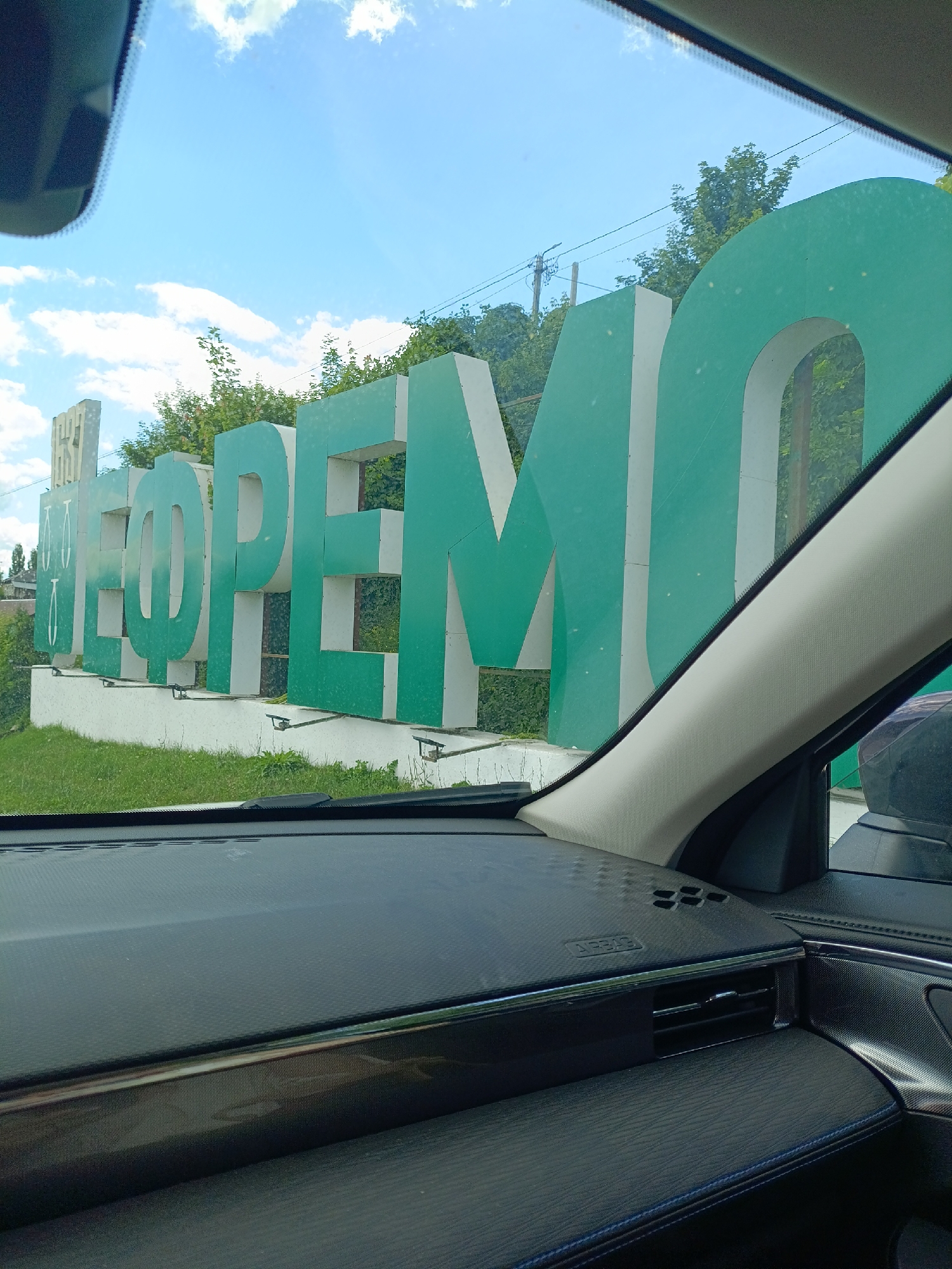 A low-angle, medium shot captures a large, three-dimensional sign that spells out "НЕФТЕМАСЛО" in green and white lettering. The sign is situated outdoors, with lush green trees and a bright blue sky with scattered clouds in the background. A glimpse of a power line and utility pole is visible behind the trees. The foreground shows the interior of a vehicle, with the dashboard and part of the windshield visible. The dashboard features a textured black surface with a glossy trim detail and an air vent. The scene appears to be a sunny day.