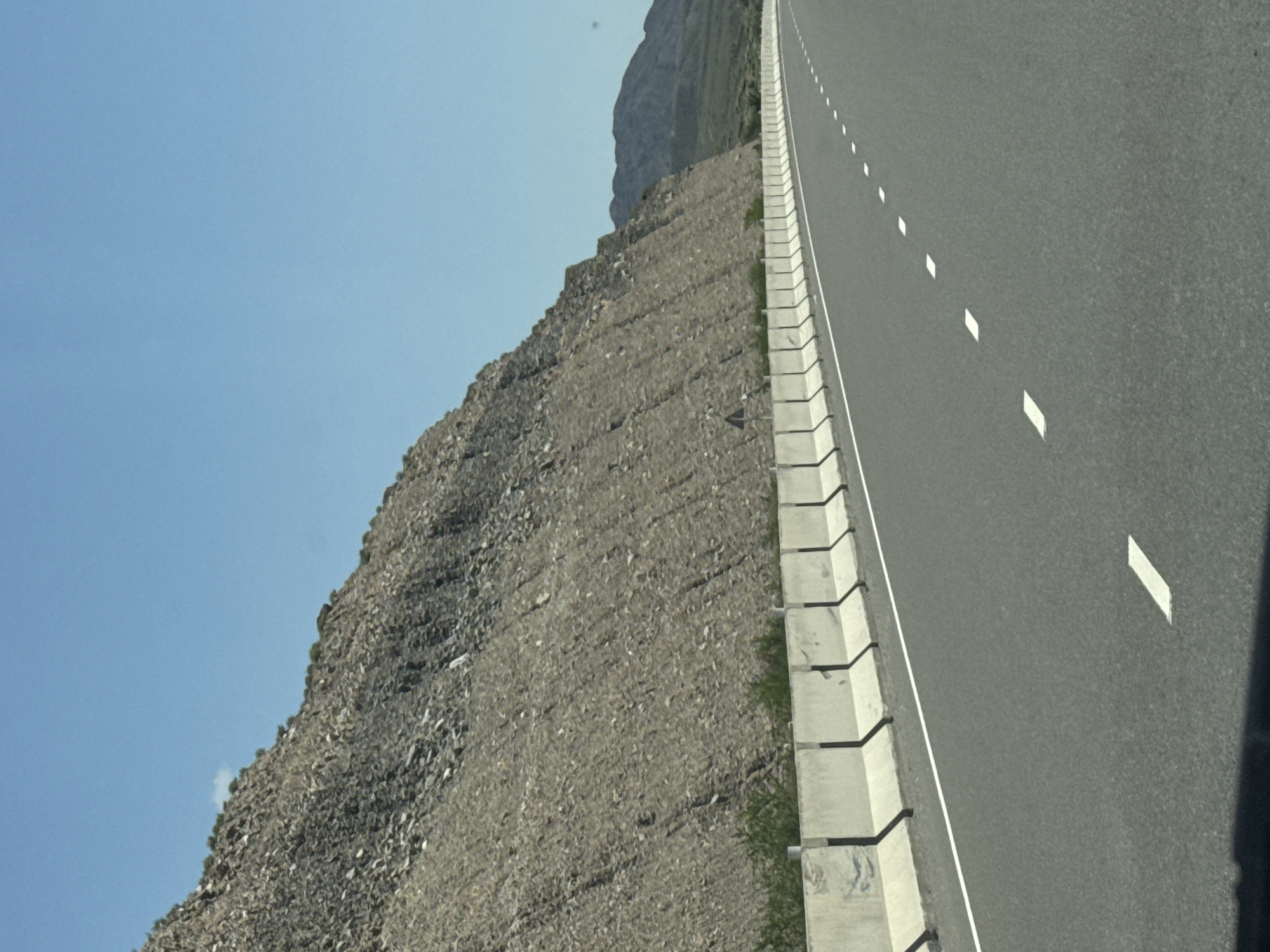 This image captures a roadside scene in the city of Kok Moynok Eki, Kyrgyzstan. The foreground features a multi-lane asphalt highway with clear white lane markings, indicating a well-maintained road. A row of precast concrete barriers runs along the right side of the highway, separating the road from a steep, rocky mountainside. The mountainside is covered in loose scree and debris, with sparse patches of green vegetation clinging to its lower slopes. The sky above is a clear, vibrant blue, with a single small white cloud visible in the upper left quadrant. In the distance, the landscape transitions to rolling hills and mountains, also appearing arid and sparsely vegetated. The overall environment suggests a highway passing through a mountainous, possibly desert-like, region.