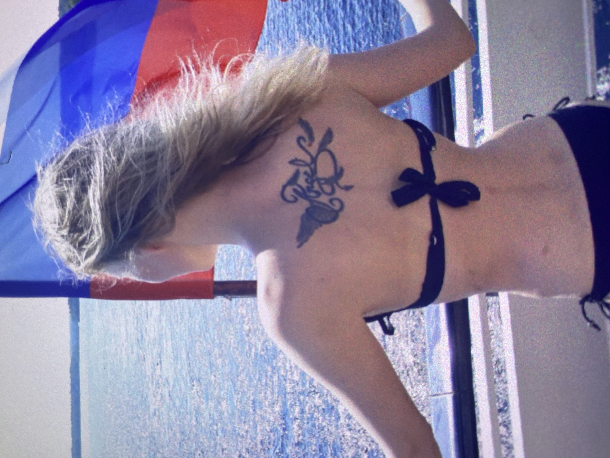 This image captures a person, likely female, from a back view, notably rotated 90 degrees clockwise. The subject has long, light-colored hair, possibly blonde or light brown, which appears somewhat textured. They are wearing a black bikini top with a bow or tie closure visible at the back, and a black bikini bottom can be partially seen. A distinct, dark-colored tattoo with an abstract design, featuring flowing lines and possibly organic or floral elements, is visible on their left shoulder blade.
The background features a body of water, possibly a lake, sea, or large pool, which exhibits a rippling texture and reflects light, indicating a bright, sunny daytime environment. In the upper left quadrant of the rotated image, a vibrant red and blue fabric object is visible, which could be part of a flag, a beach umbrella, or a canopy. On the right side, a white structure, possibly a railing or part of a building, is present.
The scene appears to be an outdoor leisure setting, perhaps by a waterfront in Saint Petersburg, Russia, given the provided context. The overall image has a slightly grainy quality, suggesting it might be an older photograph or processed with a particular filter. No visible text is present.
