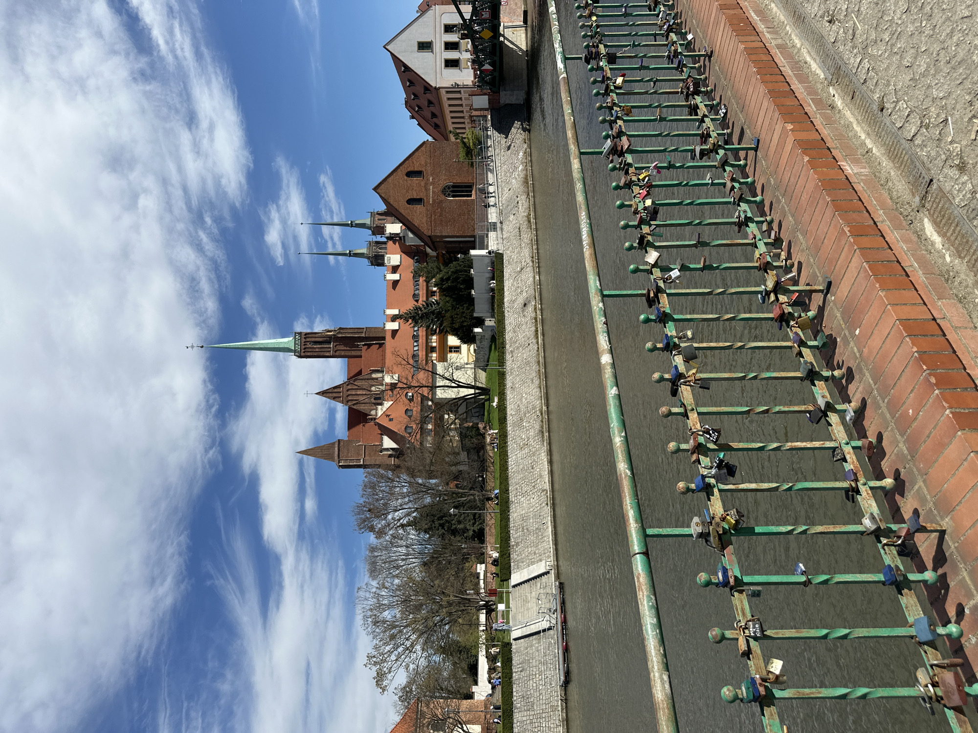 The image shows a view in Wrocław, Poland, featuring a canal lined with buildings on the left and a brick wall on the right. A green metal railing runs along the edge of the canal, adorned with numerous padlocks of various colors and sizes, presumably love locks. In the background, a cluster of buildings is visible, characterized by red brick construction and distinct architectural styles, including pointed spires. The sky is bright blue with wispy white clouds. The overall scene suggests a tourist area with a blend of historical architecture and modern expressions of affection.