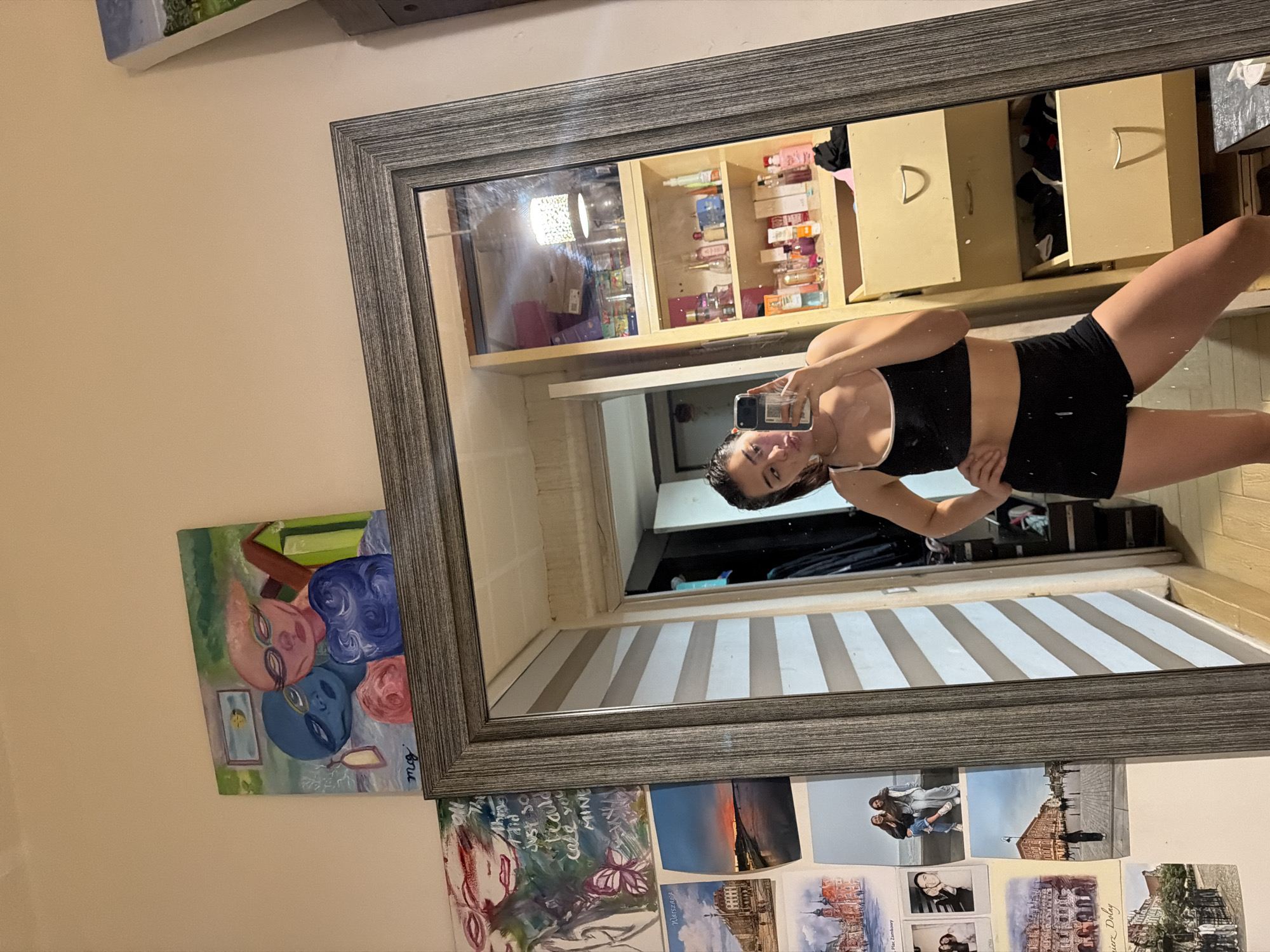 The image shows a young woman taking a mirror selfie in a room. She is wearing a black sports bra with white trim and black shorts. Her hair is pulled back in a ponytail. The mirror reflects the room behind her, which includes a closet with hanging clothes, shelves filled with various bottles, and two drawers. The walls are decorated with several framed pictures and paintings, including a painting of two figures with glasses and a sunset over a body of water. The overall impression is that of a casual, personal space. The image does not provide any information that would allow for its location to be identified as Puebla, Mexico.