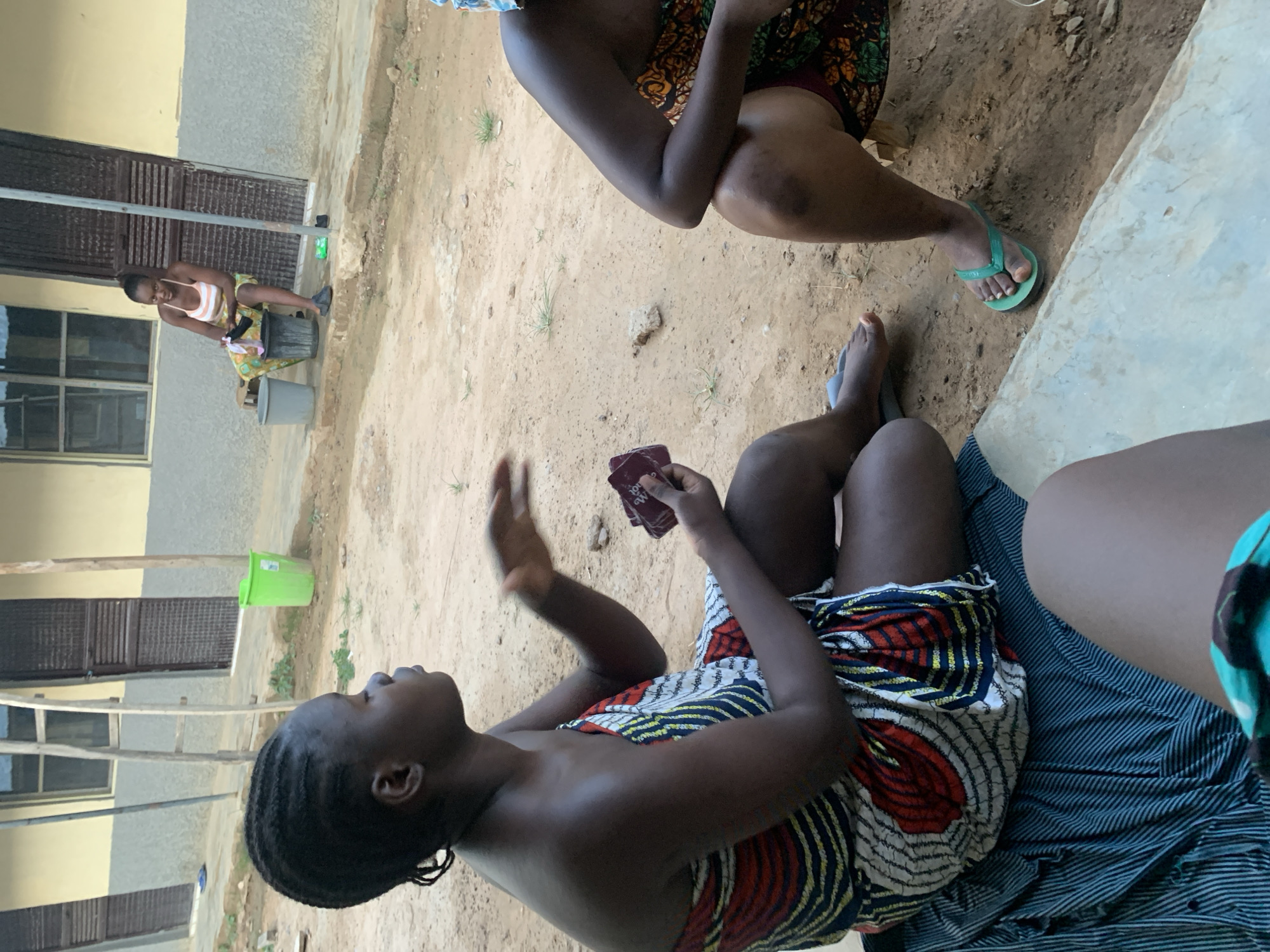This outdoor image, captured in Keffi, Nigeria, displays several individuals engaged in daily activities in a dry, unpaved communal area situated between light-colored buildings. The image is rotated 90 degrees counter-clockwise, presenting a vertical horizon.
In the foreground, a woman is seated, wearing a vibrant, patterned strapless dress and sporting dark, braided hair. She holds a small, dark object, possibly a wallet or small book, which shows partial text "MA OOL". Her right hand is raised in a gesture, suggesting she might be speaking or explaining something, as her gaze is directed upwards and slightly to her left. To her right, another individual is seated, partially visible, wearing a dark patterned fabric and a green flip-flop on one foot. A third person is mostly obscured on the far right, with only their legs and a blue and white striped fabric visible.
In the background, positioned to the left of the main group, a young woman is bent over, actively washing items in a dark bucket. She wears a striped tank top and shorts, and a faint smile is visible on her face. A grey bucket rests beside her, and a small green bin is visible further back against one of the buildings. The ground is dry and earthy, with sparse patches of grass. The buildings feature light-colored walls with dark windows and door frames. The scene takes place during bright daylight, suggesting warm, sunny weather. The overall atmosphere appears casual and communal.