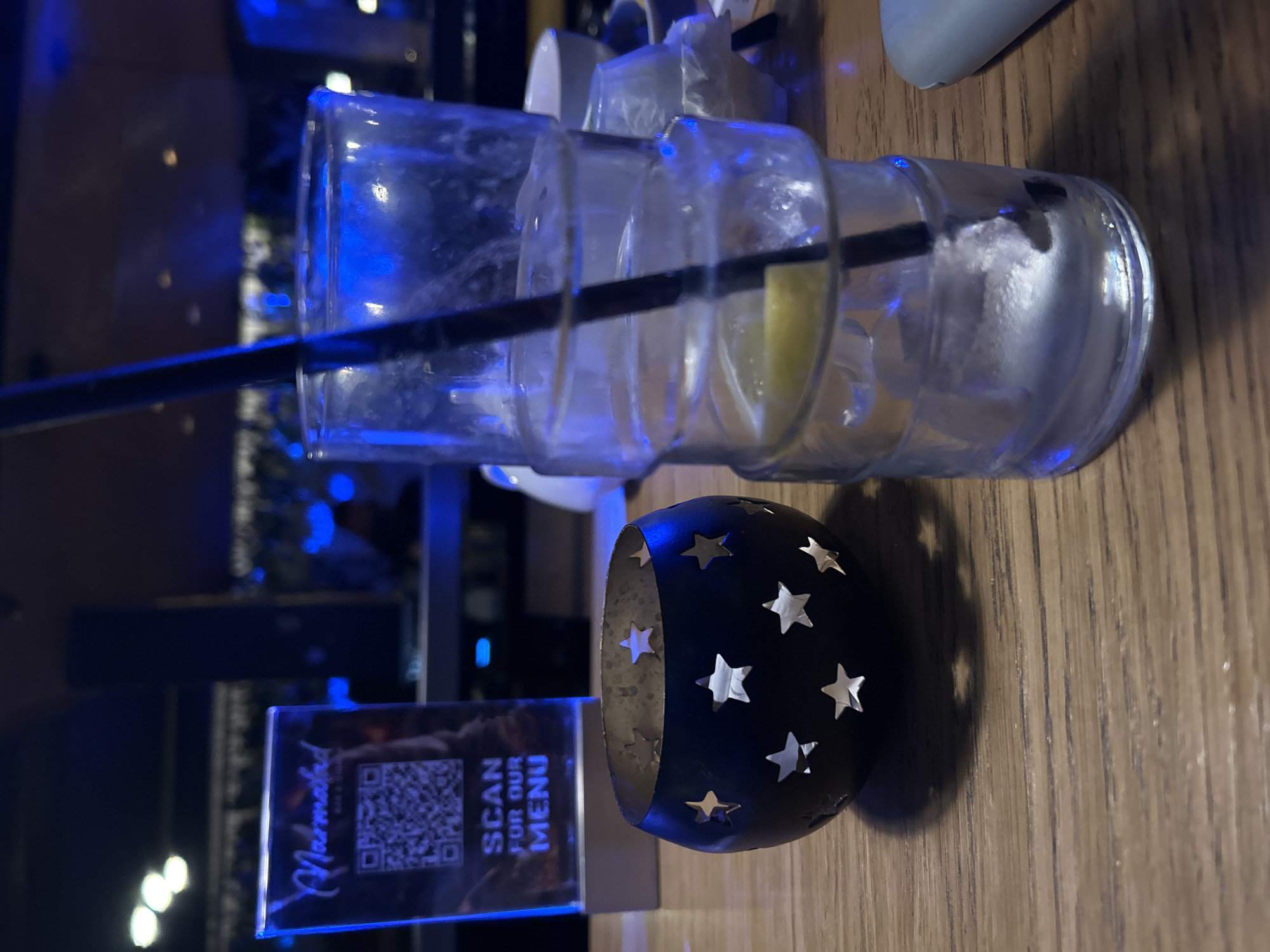 The image shows a table setting. A partially empty glass with a straw and what appears to be a lemon wedge inside is resting on its side. Next to the glass is a small, dark-colored decorative object with star-shaped cutouts. A small sign with the word "Marmalad" and "SCAN FOR OUR MENU" along with a QR code is also visible. The table appears to be made of wood. The background is dark, with indistinct lights and other elements suggesting a bar or restaurant setting. Given the sign "Marmalad" and known establishments of that name, the location is very likely a "Marmalad" restaurant in Plovdiv, Bulgaria.
