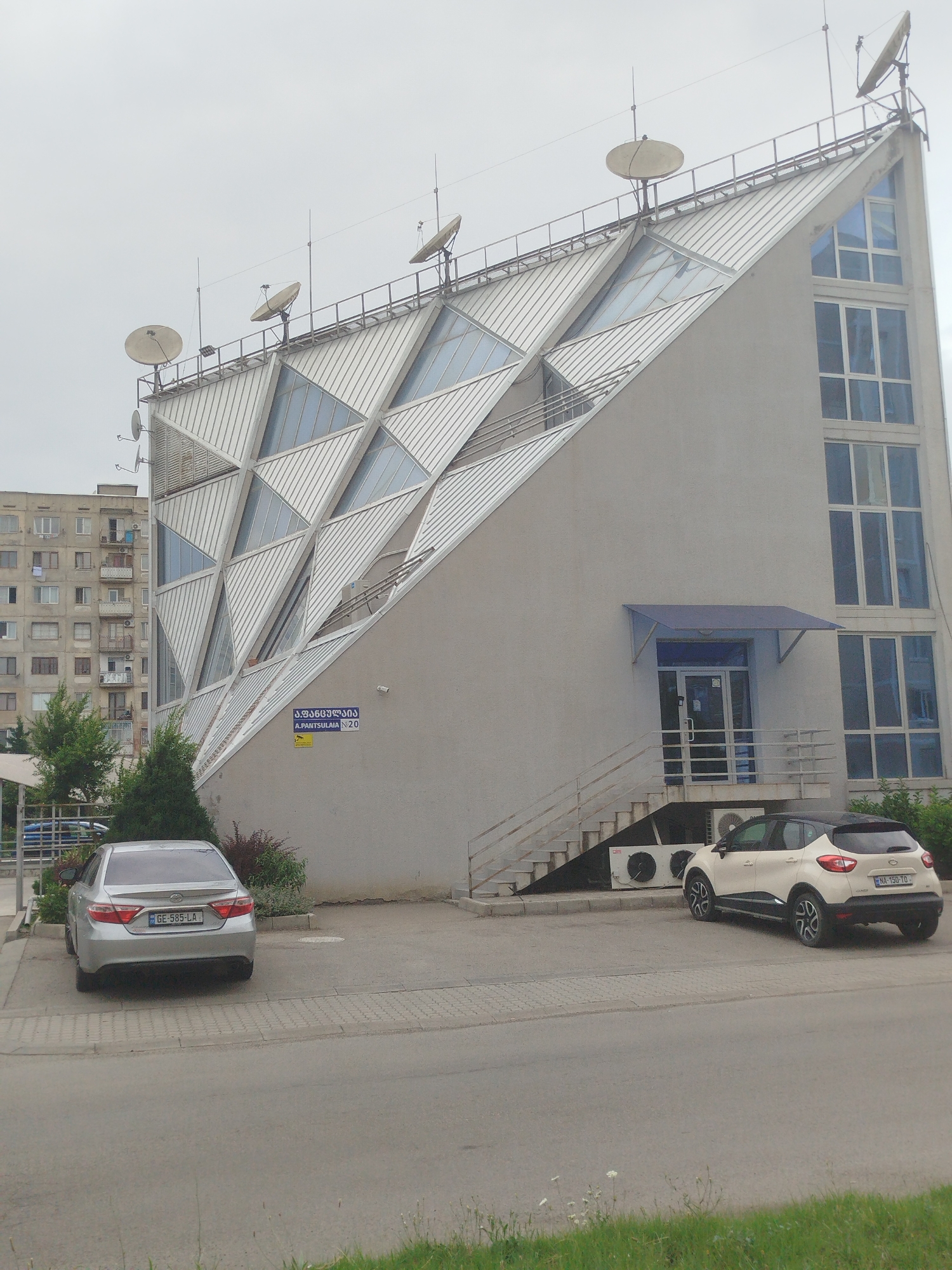 A wide shot shows the exterior of a modern, angular building with a metallic, triangular roof design in Rustavi, Georgia. The building is predominantly gray with multiple satellite dishes on the roof and a series of angled, diamond-shaped windows. To the right, the building has a section with vertical, blue-tinted windows. A blue awning covers the entrance on the right side, accessible by stairs. Two cars are parked in front of the building: a silver sedan in the foreground and a white and black crossover SUV to the right. In the background, to the left, a much older, multi-story apartment building with numerous balconies and windows is visible. The area appears to be a street or parking lot with paved surfaces and some greenery. A street sign in Georgian script is mounted on the wall of the modern building. The sky is overcast.