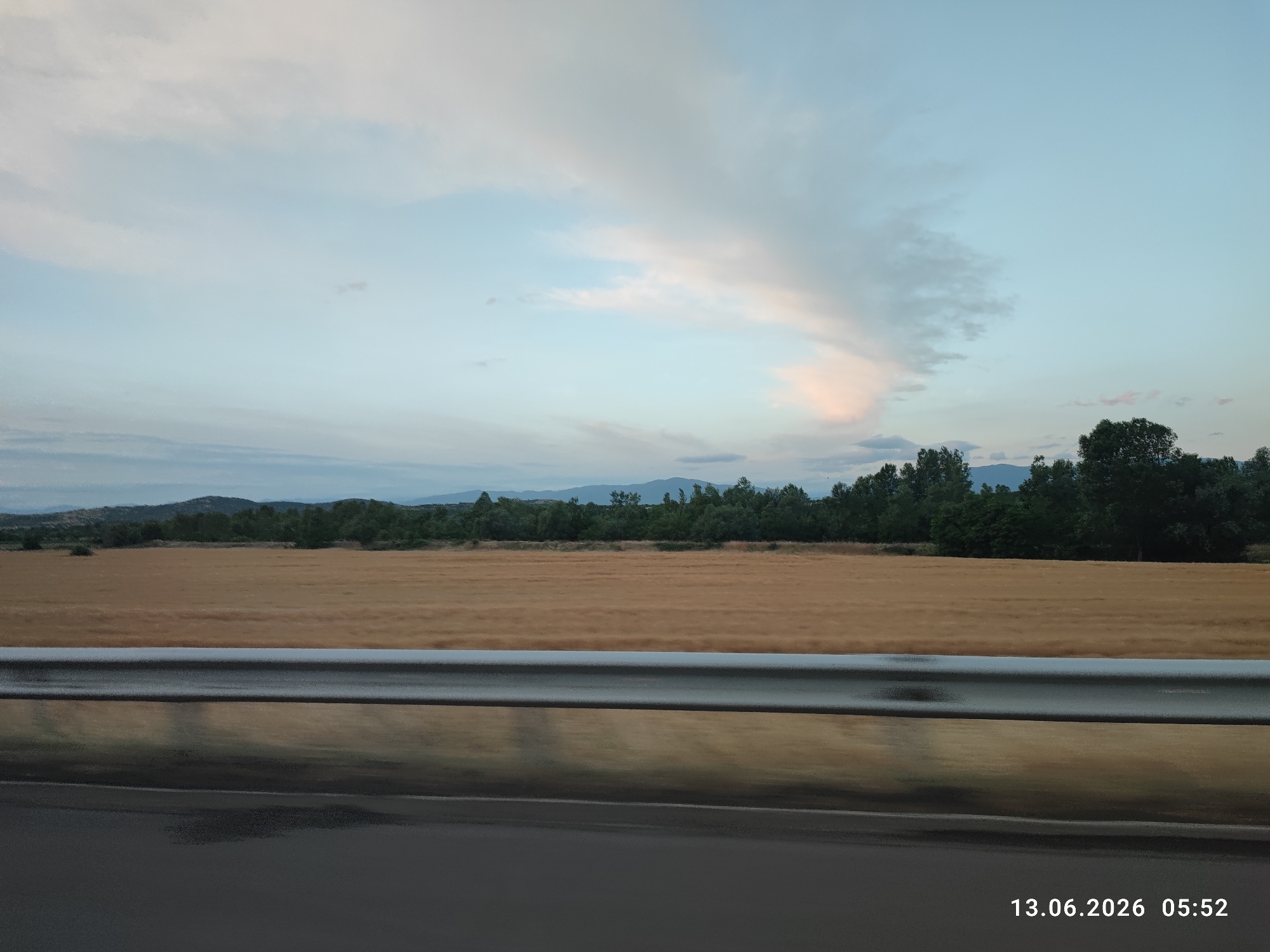 An outdoor, wide-angle, eye-level, daytime shot captures a rural roadside scene. In the foreground, a dark asphalt road transitions into a reflective metal guardrail. Beyond the guardrail, a vast field of golden crops stretches horizontally across the frame, bordered by a dense line of dark green trees. In the distance, rolling hills and mountains are visible under a pale blue sky dotted with wispy, soft clouds tinged with pink and orange hues. The timestamp "13.06.2026 05:52" is imprinted in the lower right corner. The scene appears to be located in Kalugerovo, Bulgaria.