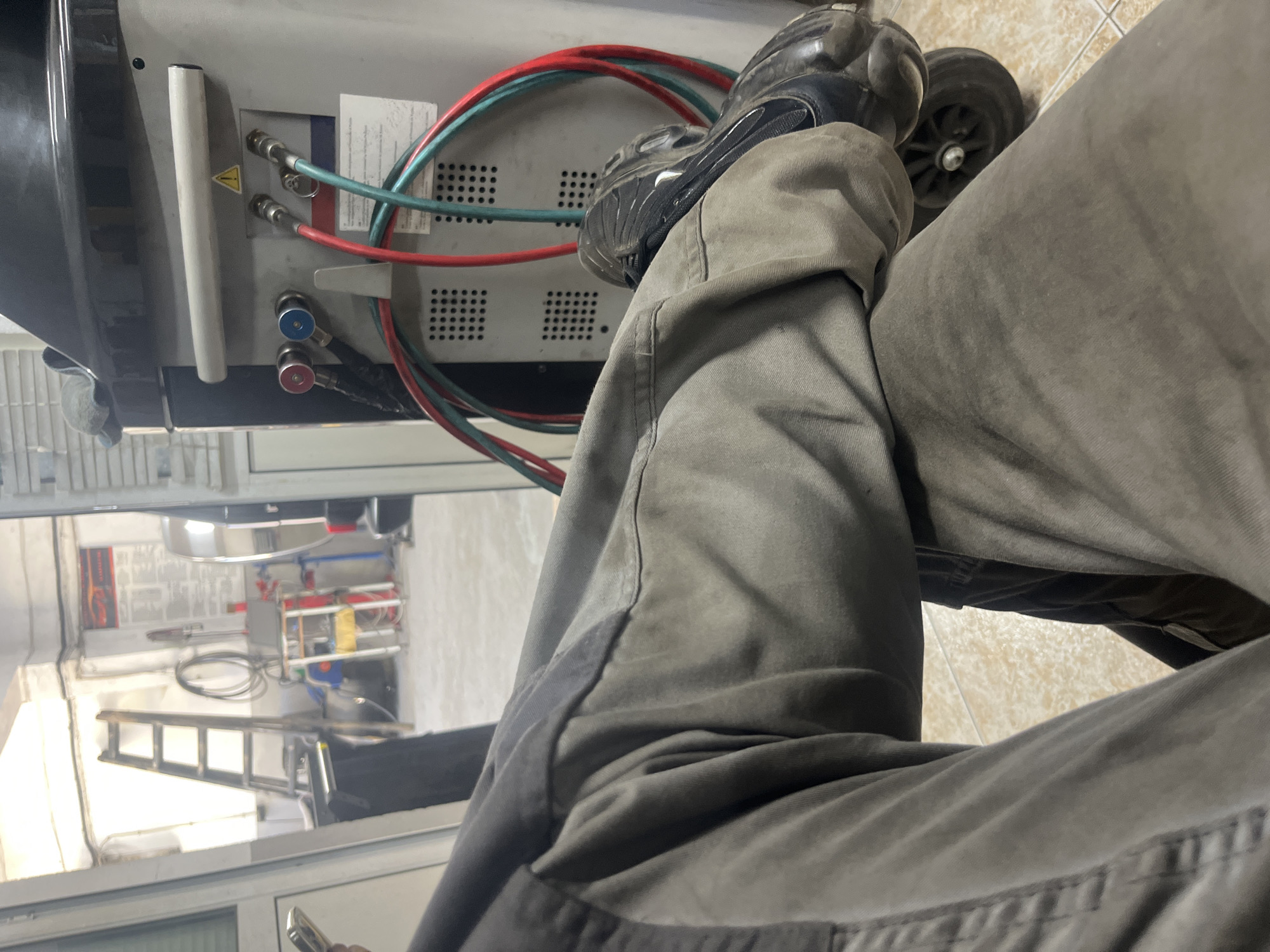 The image shows a person's legs and feet in a workshop setting. The person is wearing gray work pants and black athletic shoes. Their legs are crossed, with the left leg resting on top of the right. A portion of a piece of machinery with red and green hoses attached is visible, along with two gauges, one with a blue handle and the other with a red handle. The machinery appears to be a refrigerant recovery or charging unit, commonly used in automotive AC repair. The background shows a reflection in a window, revealing a cluttered workshop with shelves, equipment, and a ladder. The floor is tiled with a beige and white pattern. The lighting suggests an indoor environment, possibly during daytime, but no specific time is indicated. There is a yellow warning triangle sticker on the machinery. The overall scene depicts someone taking a break or waiting in a repair shop. The location context of Burgas, Bulgaria, is provided but not visually evident in the image itself.