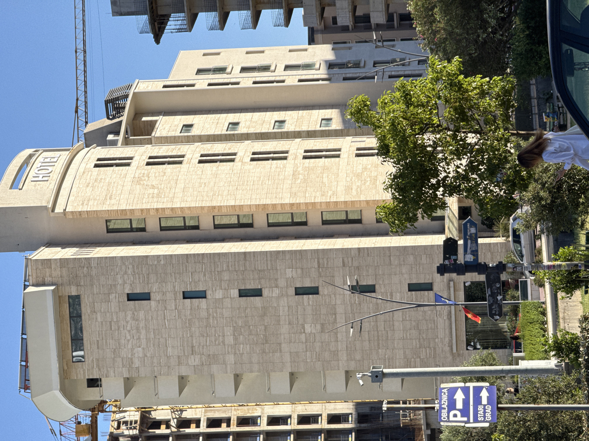 The image shows a tall, modern building, identified as a hotel due to the signage on its curved facade. The building has multiple floors with regular window placement and a beige stone facade. Construction is evident nearby, with cranes and unfinished structures partially visible. Street furniture including traffic signals and signage is in the foreground. The sign appears to show "OBILAZNICA" and "STARI GRAD", indicating a bypass and the old town.