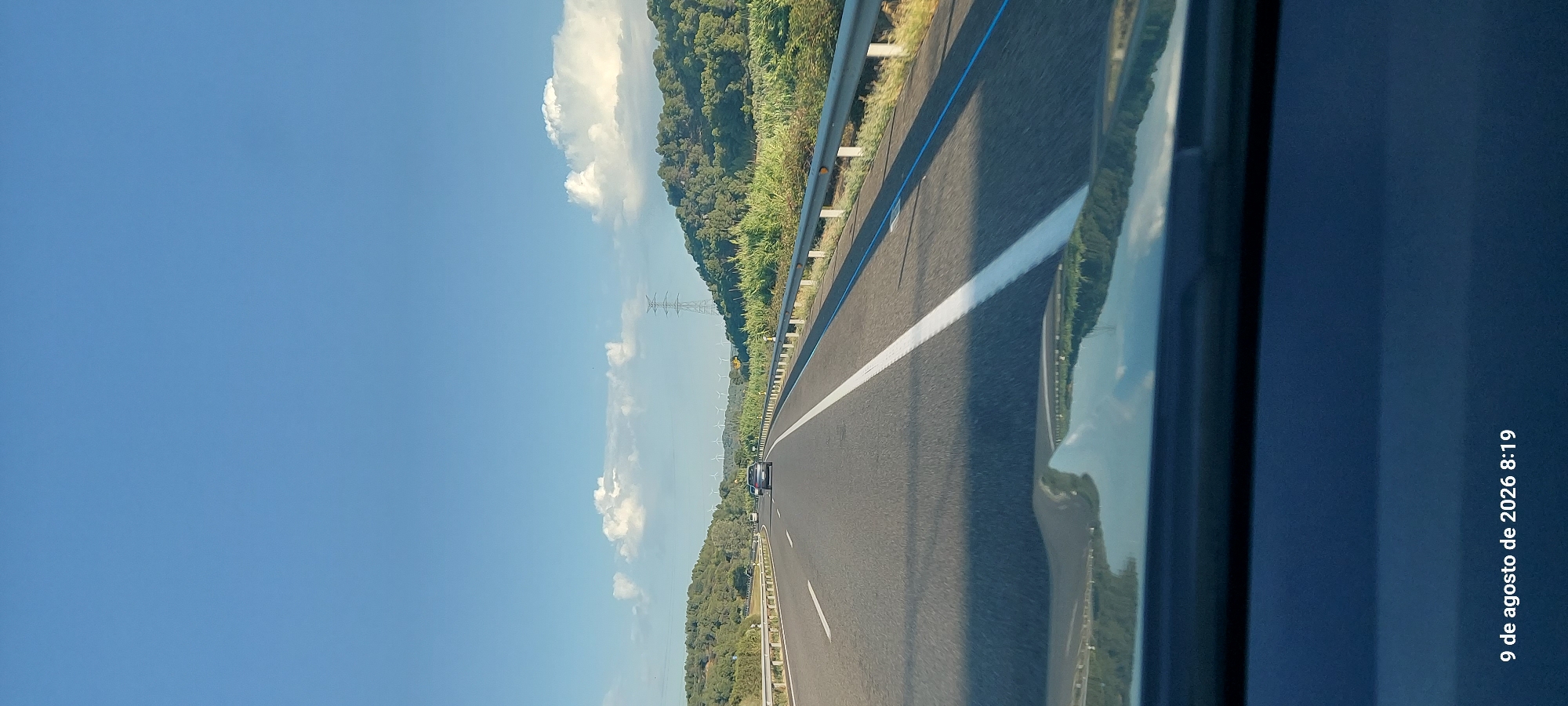 A wide-angle shot captures a highway under a clear blue sky with scattered clouds. The road stretches into the distance, flanked by guardrails and lush green vegetation. On the left, a forested hill rises, while on the right, a similar hillside is covered in dense greenery and tall grass. In the background, a few wind turbines are visible, suggesting a rural or semi-rural setting. A single vehicle is driving ahead on the highway. The foreground shows the reflection of the sky and landscape on the car's hood. The timestamp "9 de agosto de 2026 8:19" is visible in the bottom left corner. The location is identified as l'Espluga de Francolí, Spain.