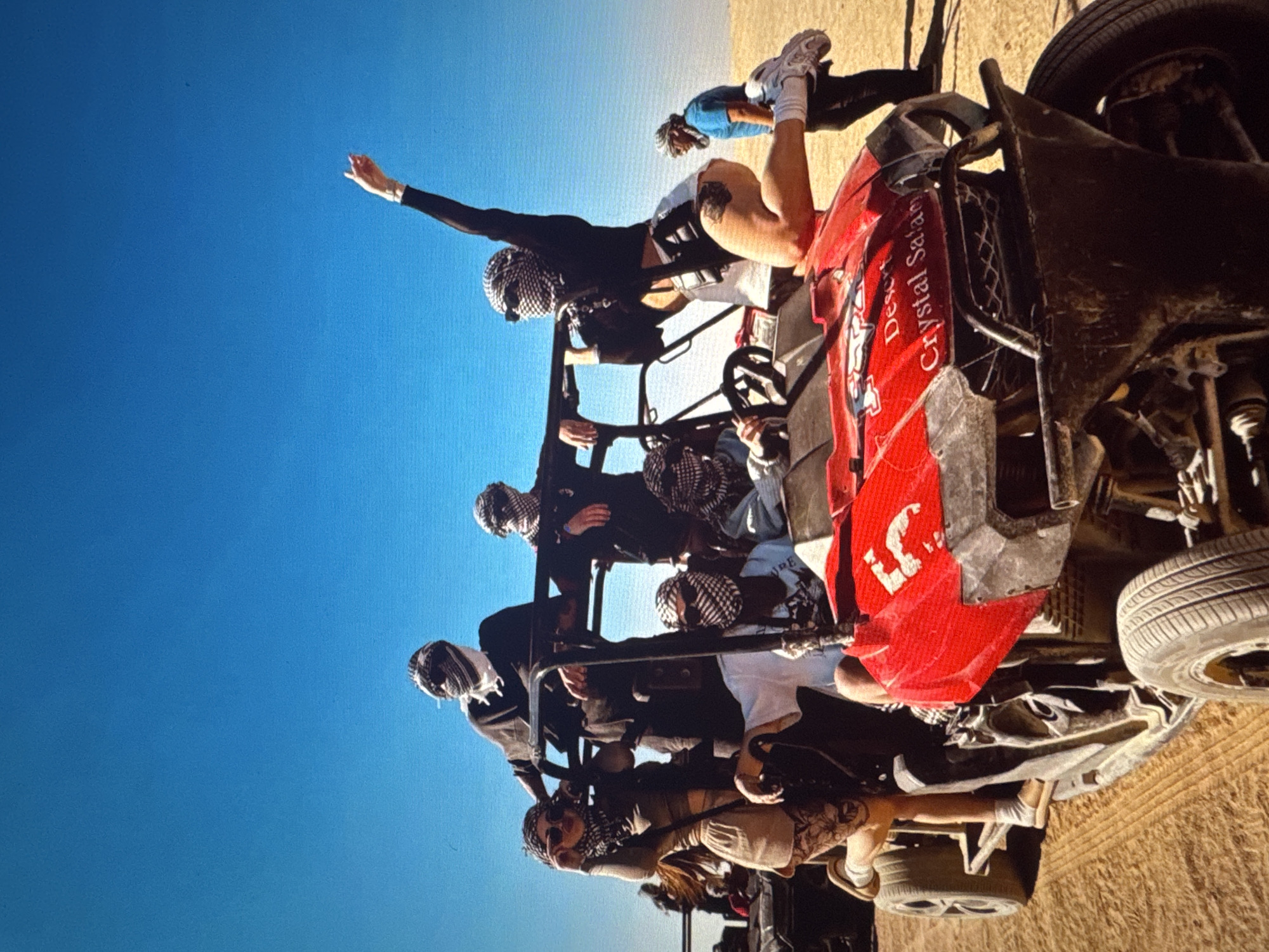 A group of people are posing in and around a red ATV in a desert environment under a clear blue sky. Several individuals are wearing keffiyeh scarves covering their heads and faces. One woman is perched on the front of the ATV, with her arm raised. Another woman is leaning on the side of the ATV, wearing sunglasses and a light-colored outfit. Two men are seated in the ATV, one with a white t-shirt and the other in a black shirt. Another man in a blue shirt and sunglasses stands in the background to the right. The ATV has "Desert Crystal Safari" written on its front. The ground is sandy and appears to be a desert landscape with tire tracks visible. The lighting suggests it is daytime with bright sunlight. This scene does not appear to be located in Tallinn, Estonia.