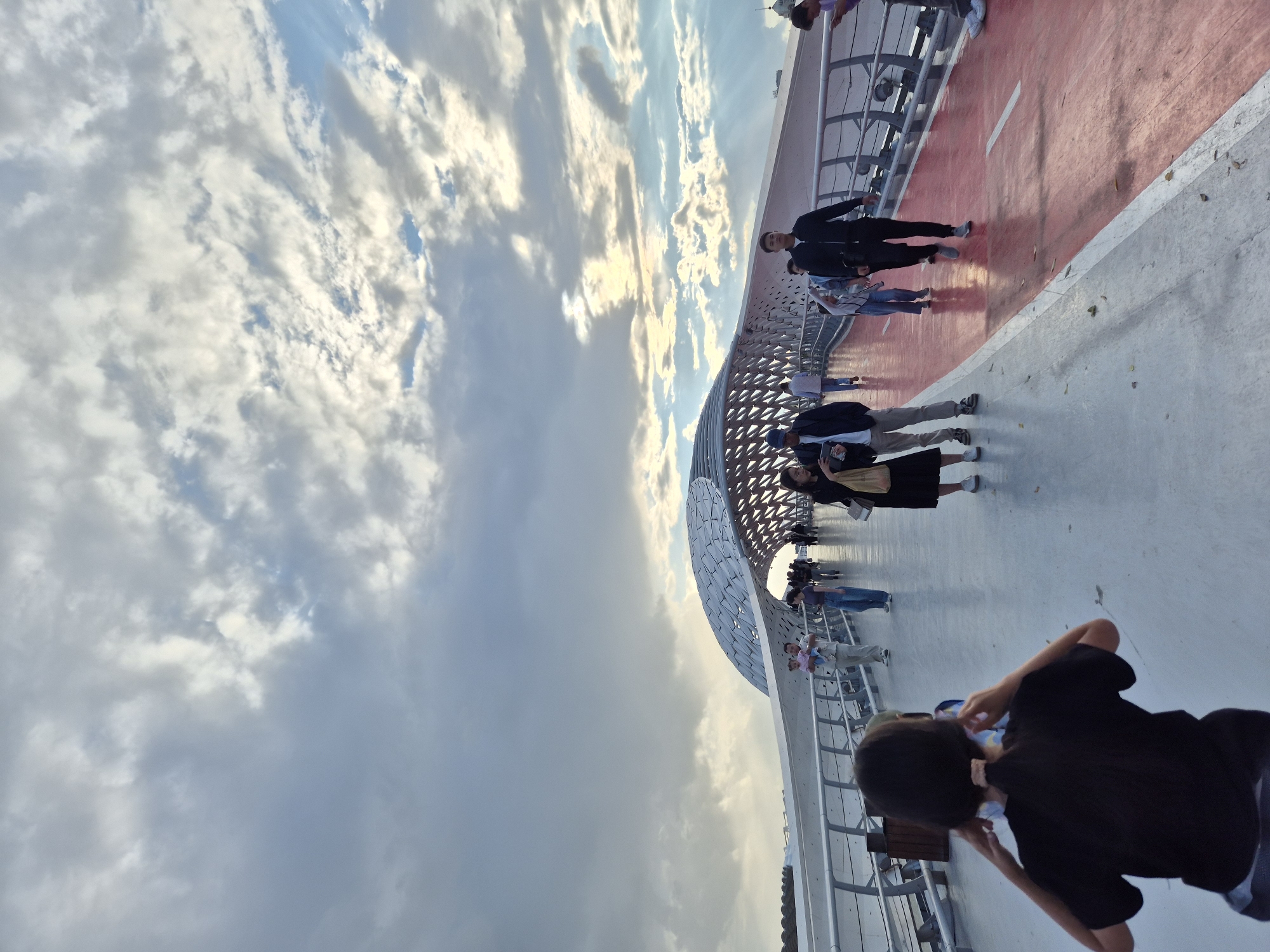 A group of people are walking across a pedestrian bridge with a unique, curved, white canopy structure overhead. The sky is filled with dramatic clouds, with shafts of sunlight breaking through in some areas, suggesting either sunrise or sunset. The bridge has a red-colored pathway marked for cycling or running, alongside a wider white pedestrian area. Several individuals are visible, some walking in groups, others alone. In the foreground, a person is facing away from the camera, appearing to adjust something near their ears, possibly headphones. The architectural style of the bridge is modern and futuristic. This bridge is the "Nur-Astana" pedestrian bridge, a notable landmark in Astana, Kazakhstan, known for its distinctive design that resembles an open palm or a blooming flower. The time of day appears to be late afternoon or early evening, given the lighting conditions.