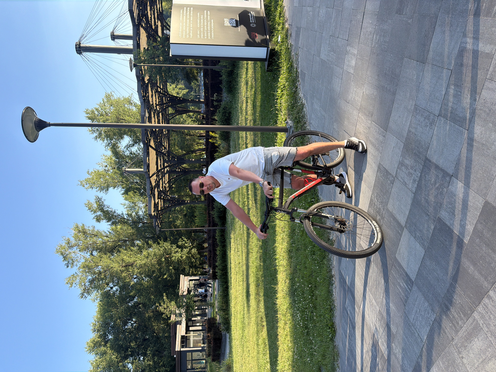 The image depicts a man in his late 30s or early 40s, wearing a white t-shirt and grey shorts, riding a bicycle on a paved walkway. He is wearing sunglasses and has a relaxed posture. The bicycle is a red and black mountain bike. The walkway is made of grey interlocking pavers. To the left of the walkway is a well-maintained lawn with scattered white flowers, and beyond that, trees and some buildings. To the right of the walkway, there is a large informational display board. In the background, there are more trees, a large modern canopy structure with a distinctive patterned roof, and a prominent lamppost. Further in the background, there are tall pylons with suspension cables, suggesting the presence of a bridge or modern infrastructure. The sky is clear and blue, and the scene is illuminated by natural sunlight, casting long shadows. This location is likely Hydropark in Kyiv, Ukraine, known for its recreational areas and modern architectural elements.