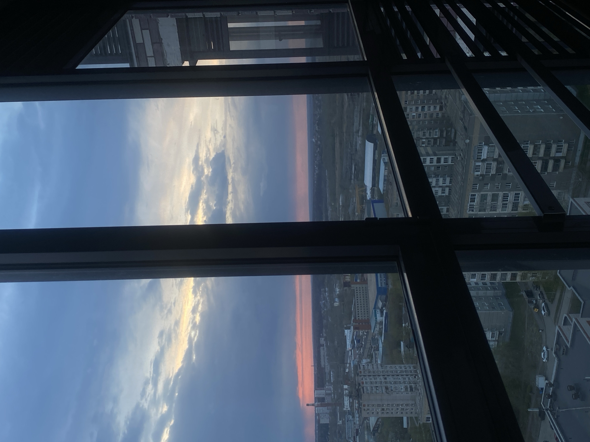 The image depicts a view from a high vantage point, likely within a tall building, in the city of Izhevsk, Russia. The scene is framed by a multi-paned window with dark framing. The view is a mix of sky and city. The sky features a blue gradient with white clouds, suggesting either sunrise or sunset, with a band of orange/pink light on the horizon. The cityscape includes residential and commercial buildings of varying heights and designs. Some of the buildings are typical Soviet-era apartment blocks, while others appear more modern. The streets and yards surrounding the buildings are visible, with a few vehicles and traces of greenery. The overall environment appears to be a densely populated urban area. Part of an unfinished building is visible on the upper left.