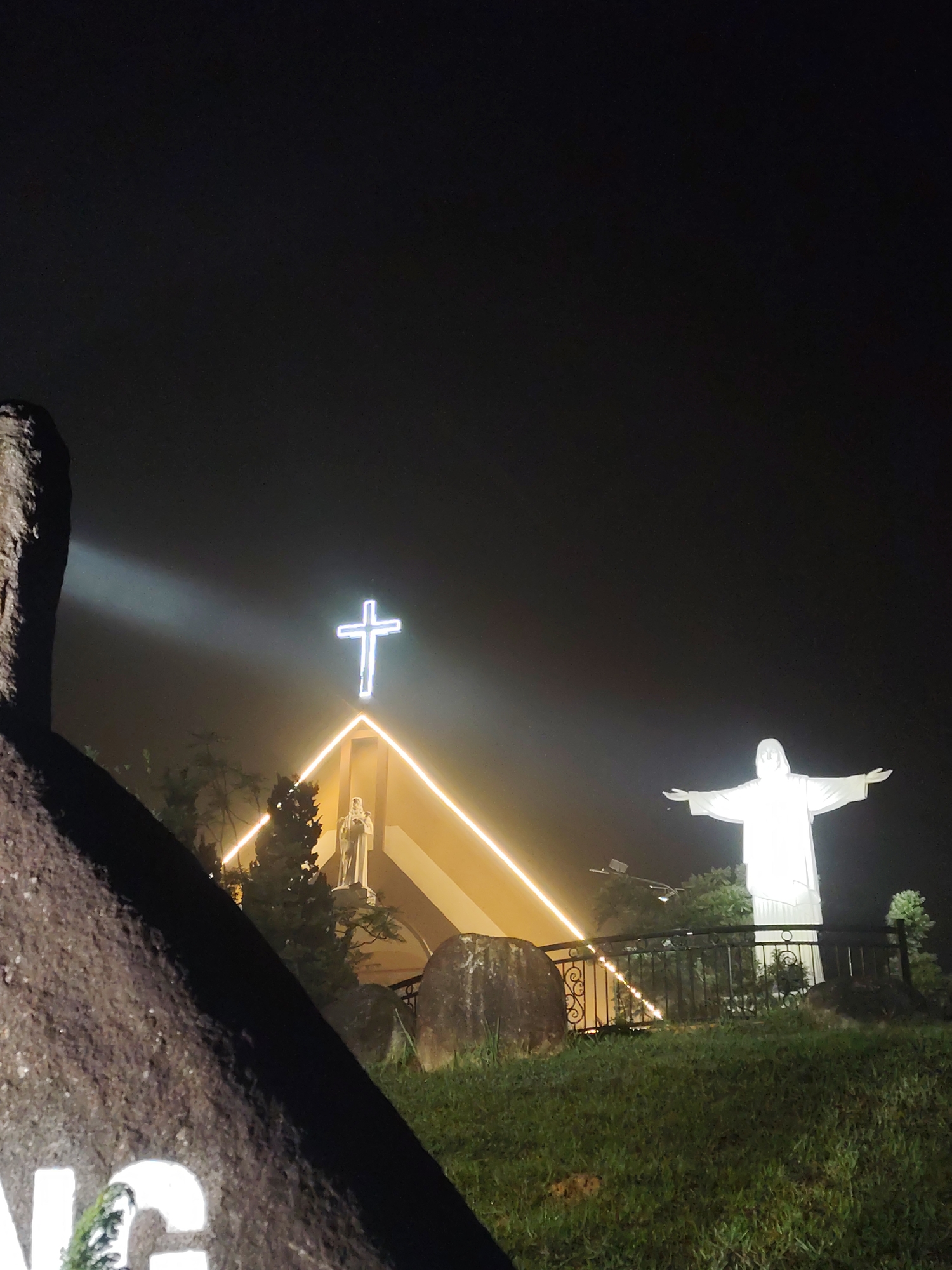 The image captures a serene nighttime scene of a religious site, likely in the misty mountainous region of Lang Biang, Da Lat, Vietnam. The foreground features a large, dark, textured rock on the left, partially obscuring the view, with visible white lettering spelling "NG" and part of another character. The ground beneath is covered in grass, interspersed with smaller rocks.
In the midground, illuminated religious structures stand prominently against a dark, atmospheric sky. To the left, a modern-styled building, possibly a chapel or church, is outlined with warm yellow neon strip lighting along its triangular roofline. A bright, glowing white cross sits atop the building, and beneath it, a smaller white statue of a robed figure, possibly a saint, is visible.
To the right, a large, radiant white statue of Christ, with outstretched arms reminiscent of Christ the Redeemer, dominates the right side of the frame. It stands on an elevated section of the terrain, in front of a black ornate metal fence or railing. Scattered trees and shrubs are visible around the structures.
A strong beam of white light emanates from the far left, cutting across the dark sky and highlighting the misty or foggy atmosphere. The overall scene is defined by the stark contrast between the luminous white statues and cross, the warm glow of the building's lighting, and the deep, dark night sky. The visible mist suggests a cool, damp evening. The scene evokes a sense of peace and spiritual solemnity, presenting a tranquil view of a sacred landmark. No specific activity is taking place.