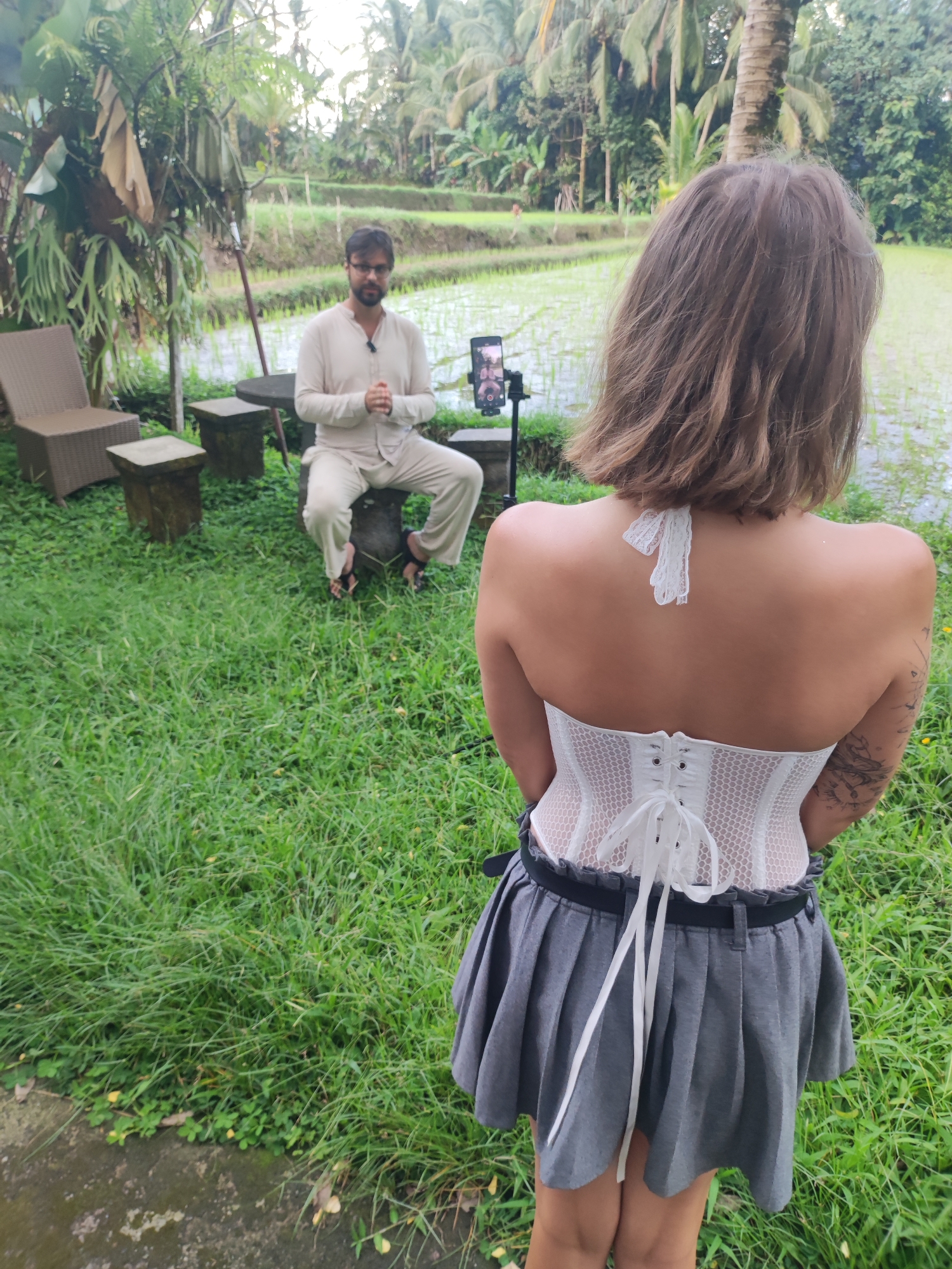 The image shows a man and a woman outdoors in a grassy area, with a rice paddy visible in the background, likely in the Tegallalang, Indonesia region. The man is sitting on a stone-like seat, wearing light-colored, long-sleeved attire and sandals. He is gesturing with his hands, possibly speaking. A smartphone is positioned on a stand in front of him, indicating he is likely being recorded. The woman is standing in the foreground, with her back to the viewer. She is wearing a white corset, a grey pleated skirt, and a dark belt. Her short hair is light brown. The environment includes various plants and trees, suggesting a natural setting. There are chairs and small tables near the man, indicating a designated area for seating.