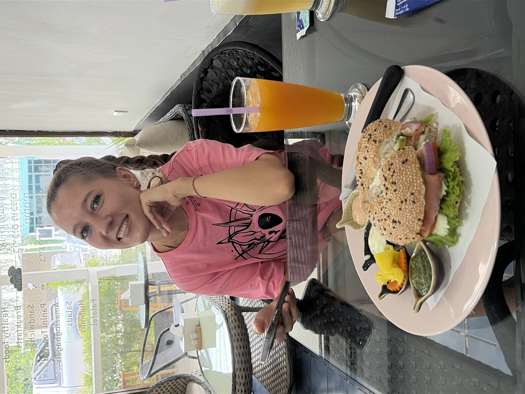 A young woman with blonde hair in braids is seated at a table in what appears to be an open-air cafe. She is wearing a pink t-shirt with a graphic print. She holds a smartphone in her left hand and is looking towards the camera with a smile, her right hand resting on her chin. In front of her on the table is a plate with a bagel sandwich, a side of pickles and carrots, and two small dishes of sauces. A glass of orange juice with a purple straw sits to her right. In the background, through a glass window, a street scene with vehicles and greenery is visible, suggesting the cafe is located in Baan Mae Nam, Thailand. The table surface is reflective, showing a distorted reflection of the scene.