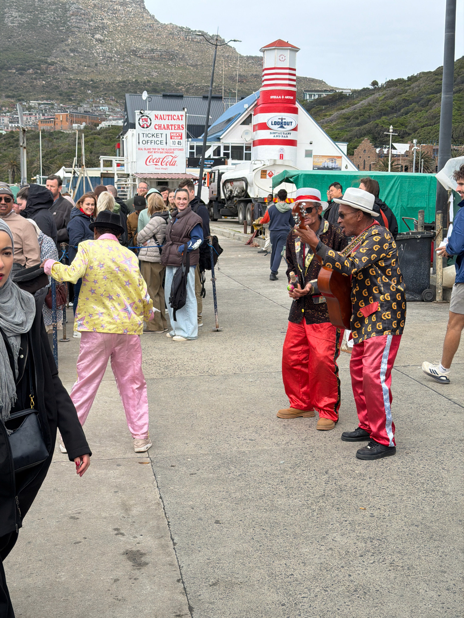 The image depicts a street scene in Hout Bay, South Africa, likely near the harbor. In the foreground, two musicians, one playing an acoustic guitar, are dressed in colorful, attention-grabbing outfits. The musician on the left wears a white fedora, sunglasses, a black jacket adorned with a pattern of gold rings, and bright red pants. The musician on the right sports a yellow jacket with a pink star pattern and matching pink pants, topped with a black fedora. They are performing for a crowd of people gathered behind them.
The background reveals a building with a distinctive red and white striped lighthouse-like structure atop it, featuring signage for "Lookout Restaurant and Bar" and "Stella Artois." Further in the background, a large truck, a Coca-Cola sign, and a "Nautical Charters" ticket office are visible. Beyond the immediate structures, a hilly landscape with scattered buildings and vegetation dominates the horizon, characteristic of Hout Bay's topography. The sky is overcast, suggesting a cloudy day. The ground is paved with concrete, showing some discoloration and debris. People in the crowd are dressed in casual attire, with varying degrees of engagement with the musicians.