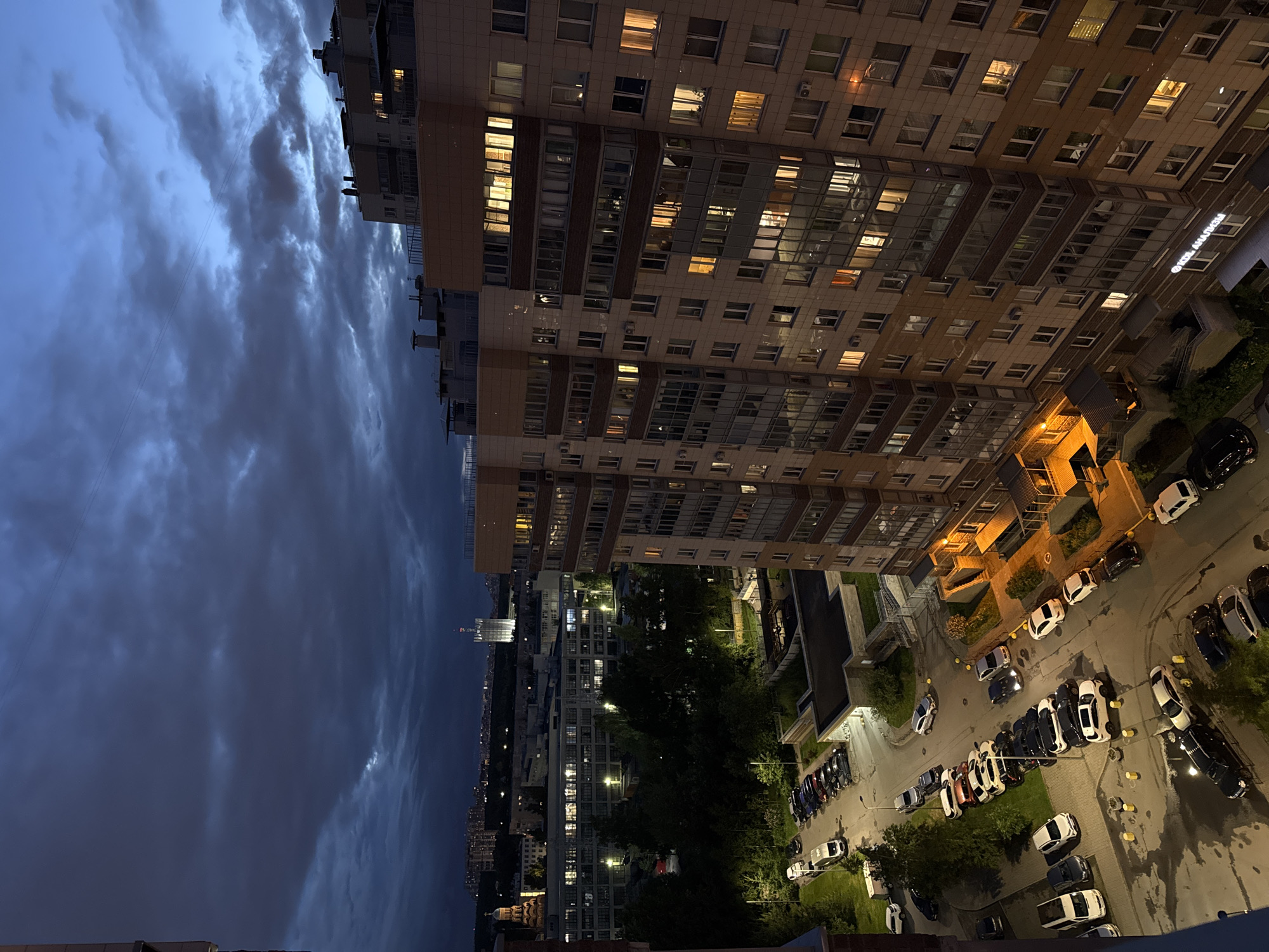 The image is an aerial view of a residential building in a city at dusk. The building is tall with multiple floors and balconies, and lights are visible in many of the windows, suggesting occupancy. Below the building, a street is lined with parked cars of various colors and models. Some of the streetlights are illuminated, casting a warm glow on the wet asphalt, indicating recent rain. Trees and other buildings are visible in the background, along with a prominent tall, illuminated structure in the distance. The sky is dark and cloudy, characteristic of twilight. The scene appears to be a typical urban environment at the end of the day.