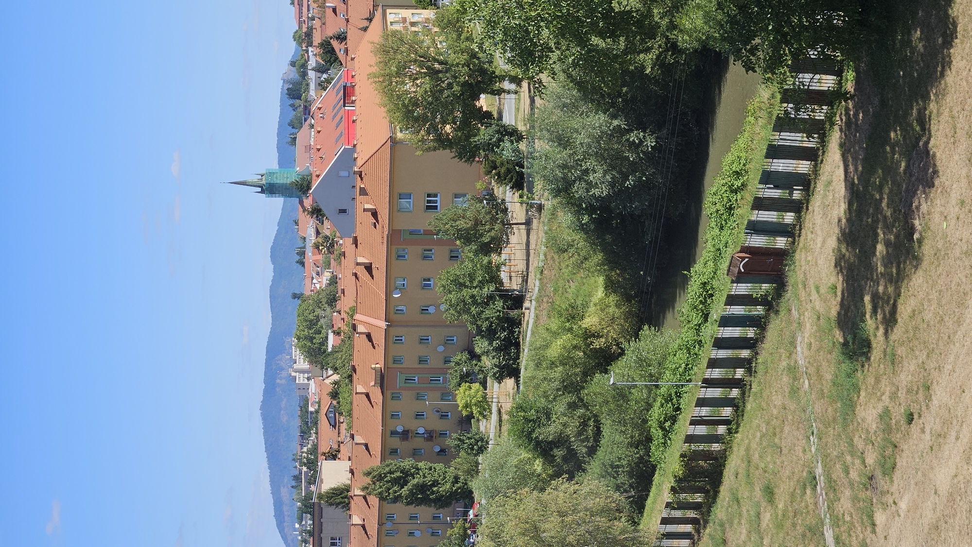 This is a vertical, eye-level, outdoor, daytime shot of a residential area in Prešov, Slovakia. In the background, a green church steeple rises above the orange-tiled roofs of apartment buildings. The middle ground features more apartment buildings and lush green trees. A river flows along the right side of the frame, bordered by a metal fence with a single brown gate on the left. The foreground consists of dry, brown grass with a dappled shadow from overhead trees. The sky is clear blue with a few wispy clouds.