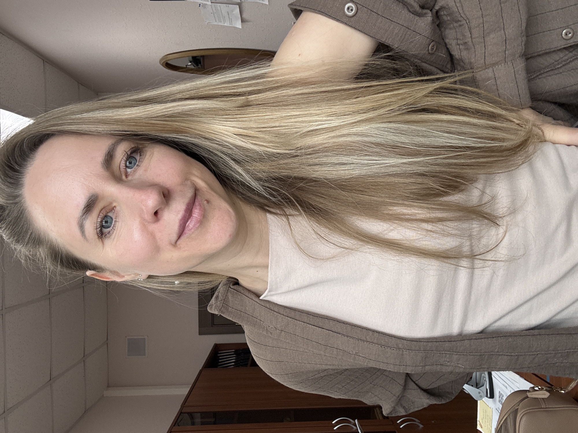 A woman with blonde hair and blue eyes is taking a selfie indoors. She is wearing a light-colored t-shirt and a brown striped shirt. Behind her is an office environment, including a ceiling with square tiles, a wooden cabinet, a mirror, and paperwork on a wall. It is not possible to determine the location in Chaikovsky, Russia from this image.