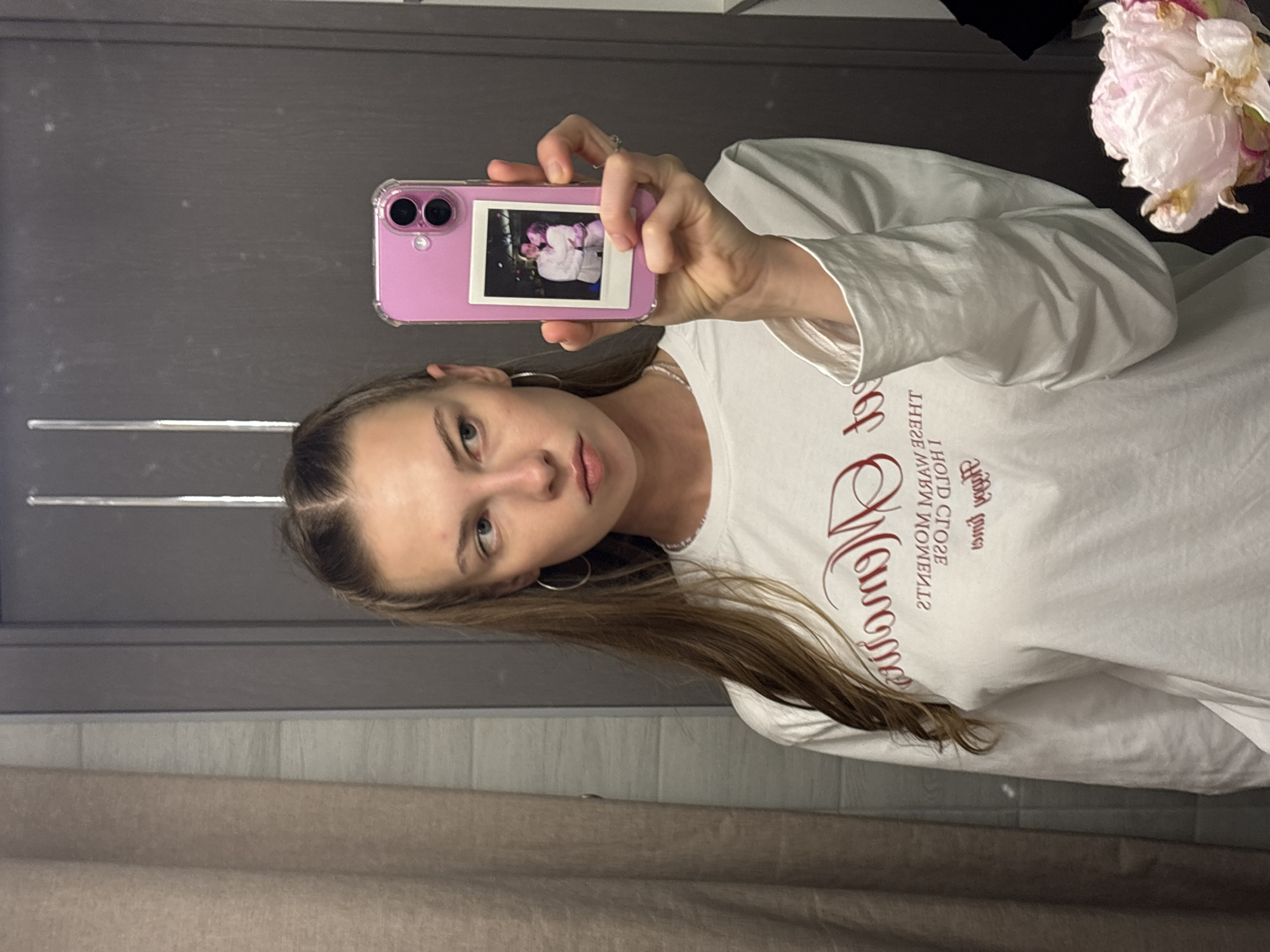 A young woman with long, brown hair, styled in two braids, takes a selfie in a bathroom. She is wearing a white long-sleeved shirt with red lettering. Her left arm is bent, and she is holding a pink phone with a clear case. A small, polaroid-style photo is attached to the back of the phone case, showing two people embracing. The woman has hoop earrings in her ears and appears to be looking at her reflection in the mirror. Behind her, a grey door is visible, and to the right, a pale pink flower with delicate petals is in the frame. The overall scene suggests a casual, personal moment captured by the woman herself. The lighting appears to be indoor, possibly artificial. The visible text on her shirt reads, in reverse due to the mirror, "OLD CLOSE MOMENTS" and a stylized word which is difficult to decipher completely.