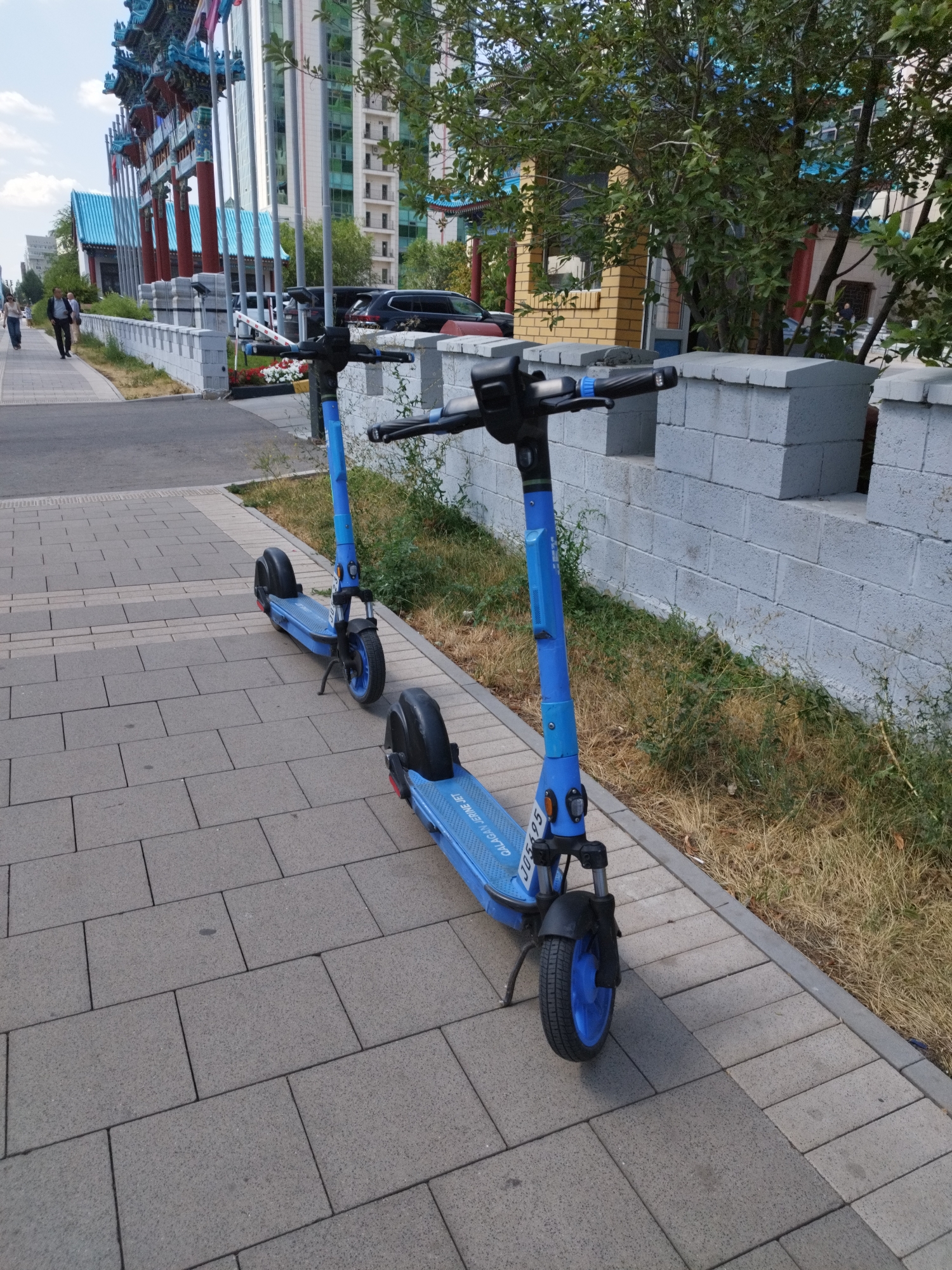 Two blue electric scooters are parked side-by-side on a paved walkway in front of a white brick wall and dry grass. The scooter in the foreground is closer to the camera, and its front wheel is turned slightly to the left. The second scooter is slightly behind and to the left of the first. In the background, a street with parked cars and people walking is visible, along with a modern building with green windows. Further back, an ornate structure with red pillars and blue roofing, typical of Astana's architecture, is present. The scene suggests an urban environment in Astana, Kazakhstan, likely a public area with shared transportation options.