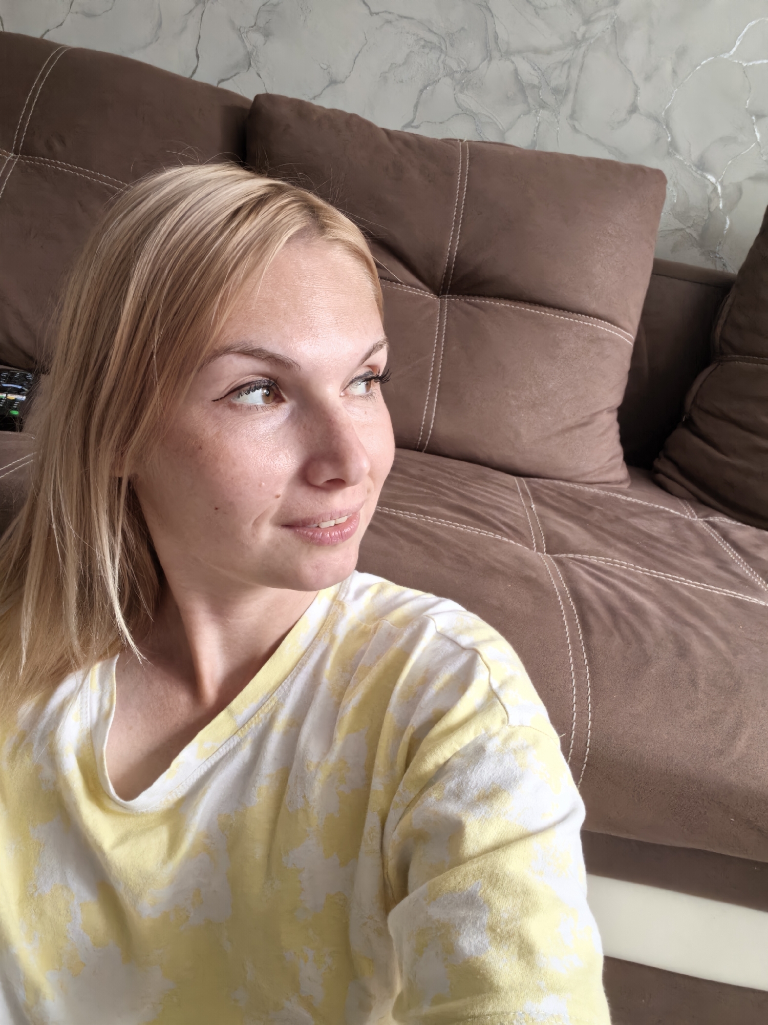 The image shows a close-up, head-and-shoulders shot of a blonde woman with fair skin, light brown eyes, and freckles. She is looking to her right, with a slight smile on her lips. She is wearing a yellow and white tie-dye t-shirt. Behind her, a brown sofa with visible stitching is visible, and a marble-patterned wall can be seen in the upper right corner. The lighting suggests an indoor setting, likely a living room. There are no visible indications of the city of Sterlitamak, Russia.