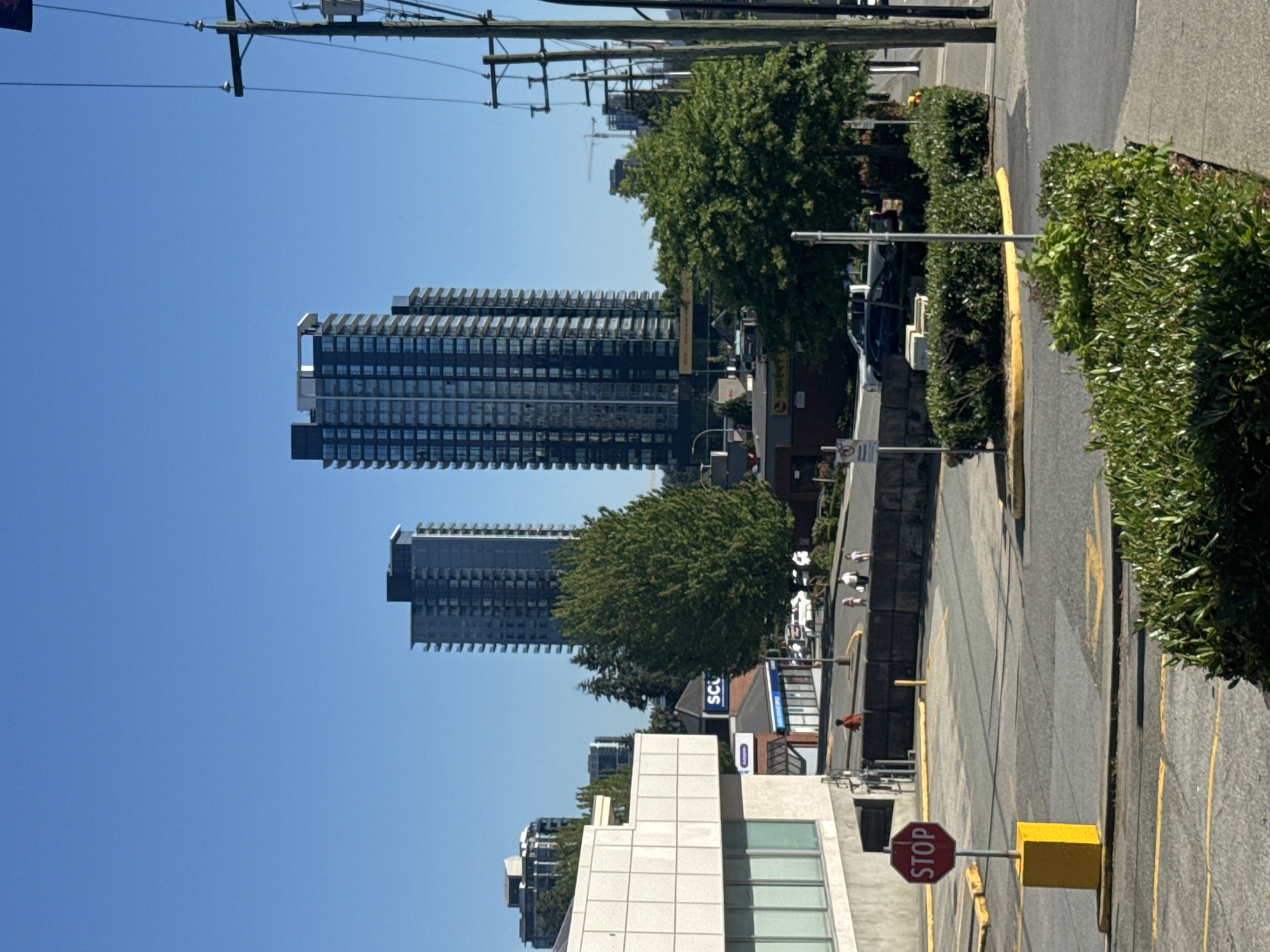 In an urban environment under a clear blue sky, a street view captures a scene in Burnaby, Canada. In the foreground, a yellow post supports a red octagonal stop sign, its white lettering distinct against the crimson background. The street is paved with asphalt, marked with yellow lines indicating parking spaces and traffic flow. Lush green bushes line the edge of a sidewalk.
Further into the scene, a light-colored, modern building with large windows is visible on the left. Across the street, a dark-colored building with an awning is situated near a mature, green tree. Beyond these structures, two tall, modern apartment buildings dominate the skyline, their glass facades reflecting the sunlight. Other buildings and trees are scattered throughout the midground and background. A few people are walking on the sidewalk and street. In the distance, construction cranes are present, indicating ongoing development. Power lines and utility poles are prominent on the right side of the image. The overall impression is one of a sunny day in a developed urban area.