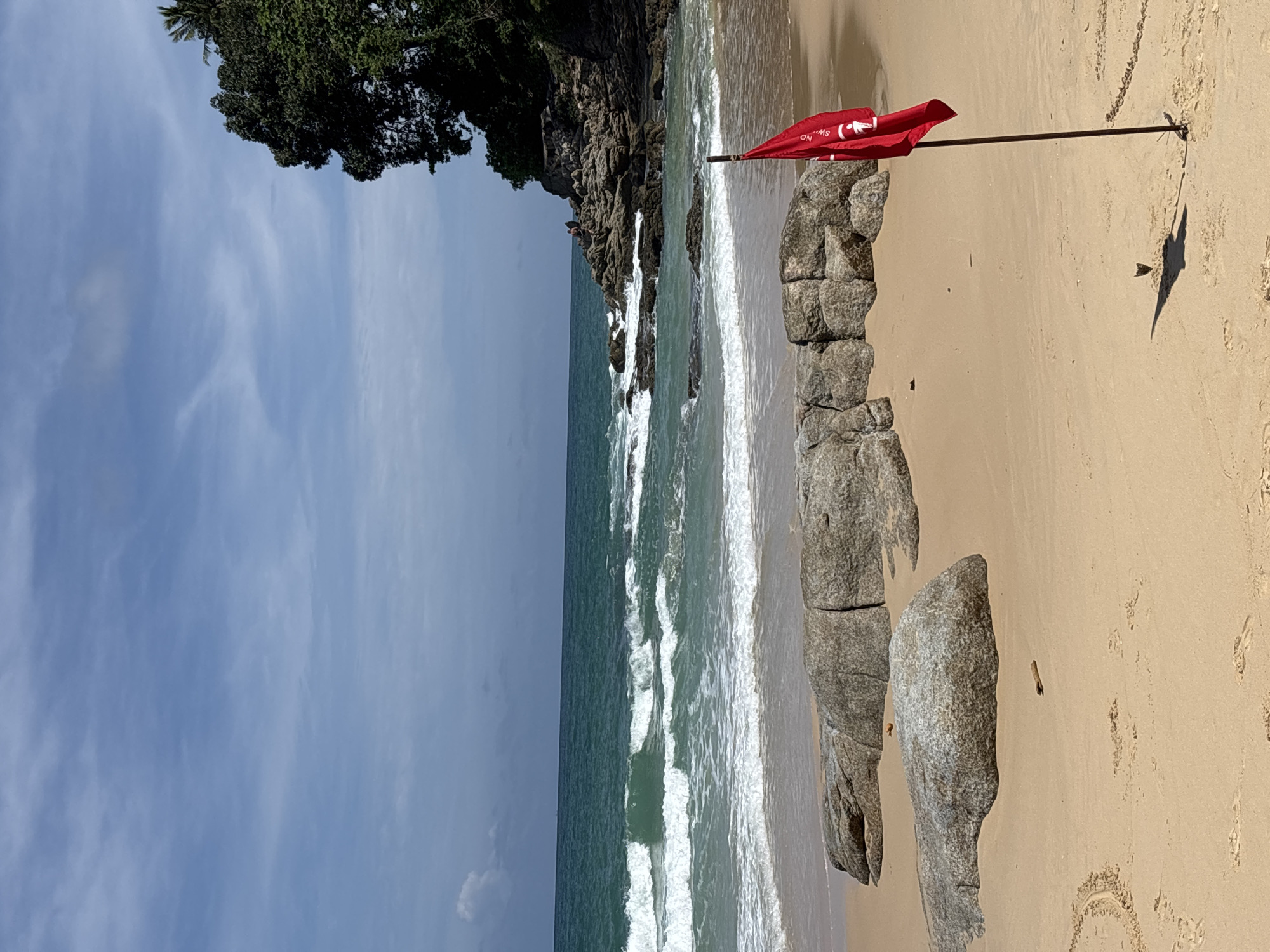 The image depicts a beach in Choeng Thale, Thailand. The sky is partly cloudy. To the left, a rocky headland with trees encroaches onto the beach. Waves are breaking along the shore. A red flag indicating “no swimming” is placed on the sand, near a rock formation. The sand is light-colored. A shadow cast by the flag and pole is visible.