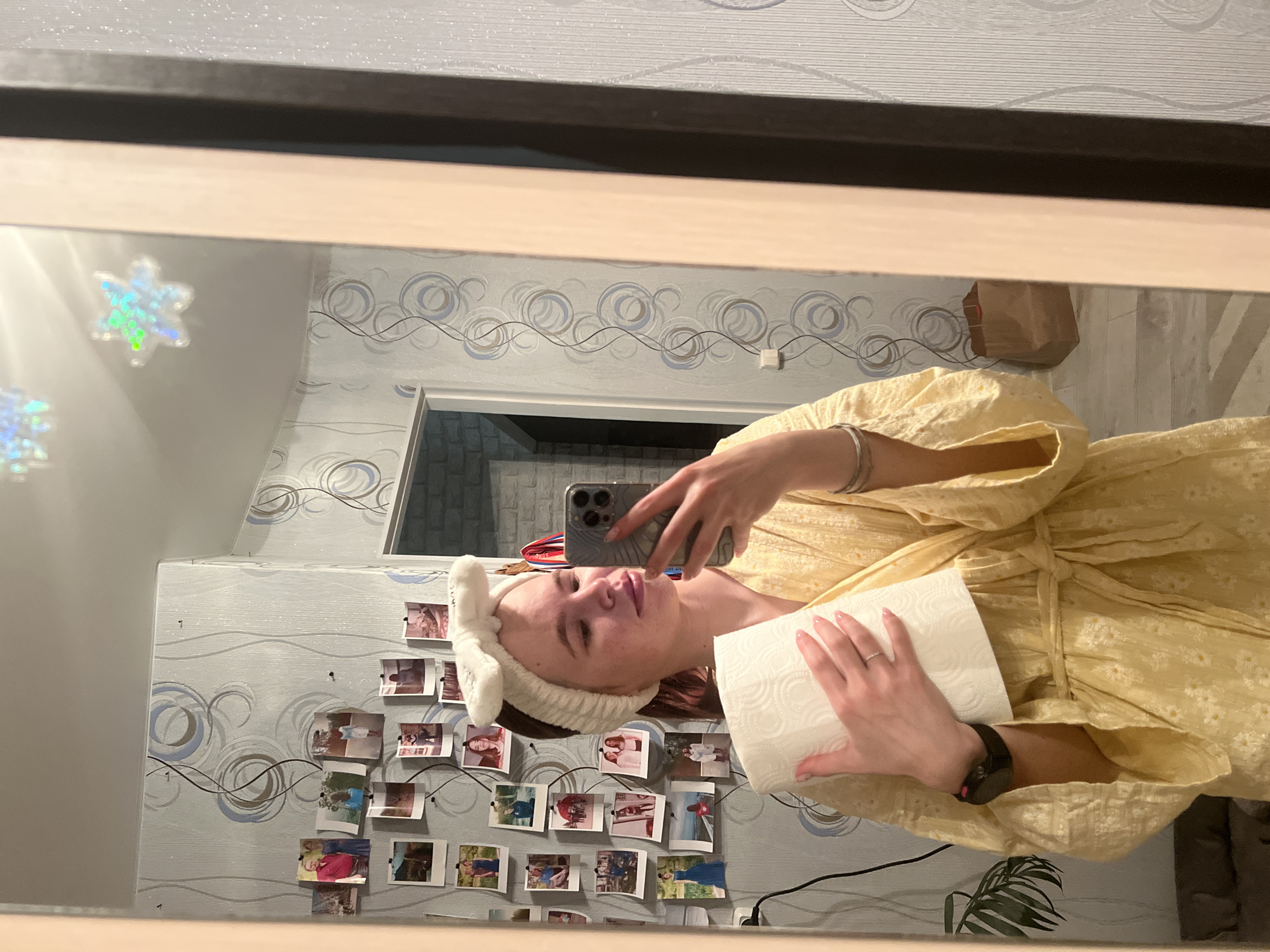 A woman is taking a selfie in a mirror. She is wearing a yellow dress and a white headband. She is holding a roll of paper towels. The background includes a wall with a swirly pattern and numerous small photos attached to it. The location is not evident from the image, although it might be related to Chaikovsky, Russia.