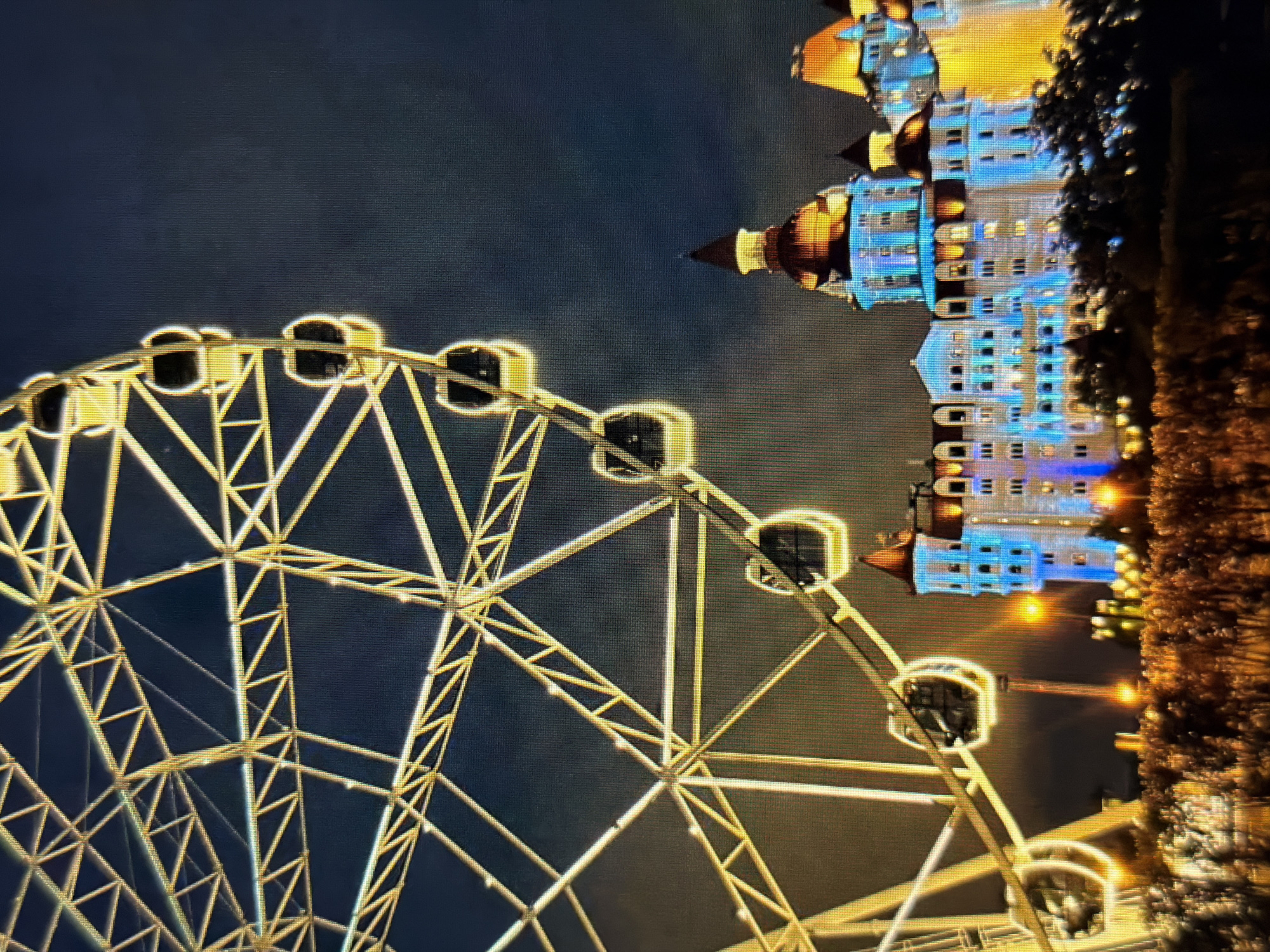 This is a night-time photograph of the Ferris wheel and the Ice Palace (Bolshoy Ice Dome) in Sirius, Russia. The Ferris wheel, illuminated with yellow lights, dominates the left side of the frame, its intricate metal structure visible against the dark sky. Several gondolas are suspended from the wheel, also outlined with light. To the right of the Ferris wheel, the Ice Palace is prominently featured, its facade lit with vibrant blue and yellow lights. The building has a distinct castle-like architecture with multiple towers and ornate roofs. The foreground consists of a blurry area of illuminated foliage and lights, suggesting a park or promenade area. The overall scene depicts an amusement park or entertainment complex at night.