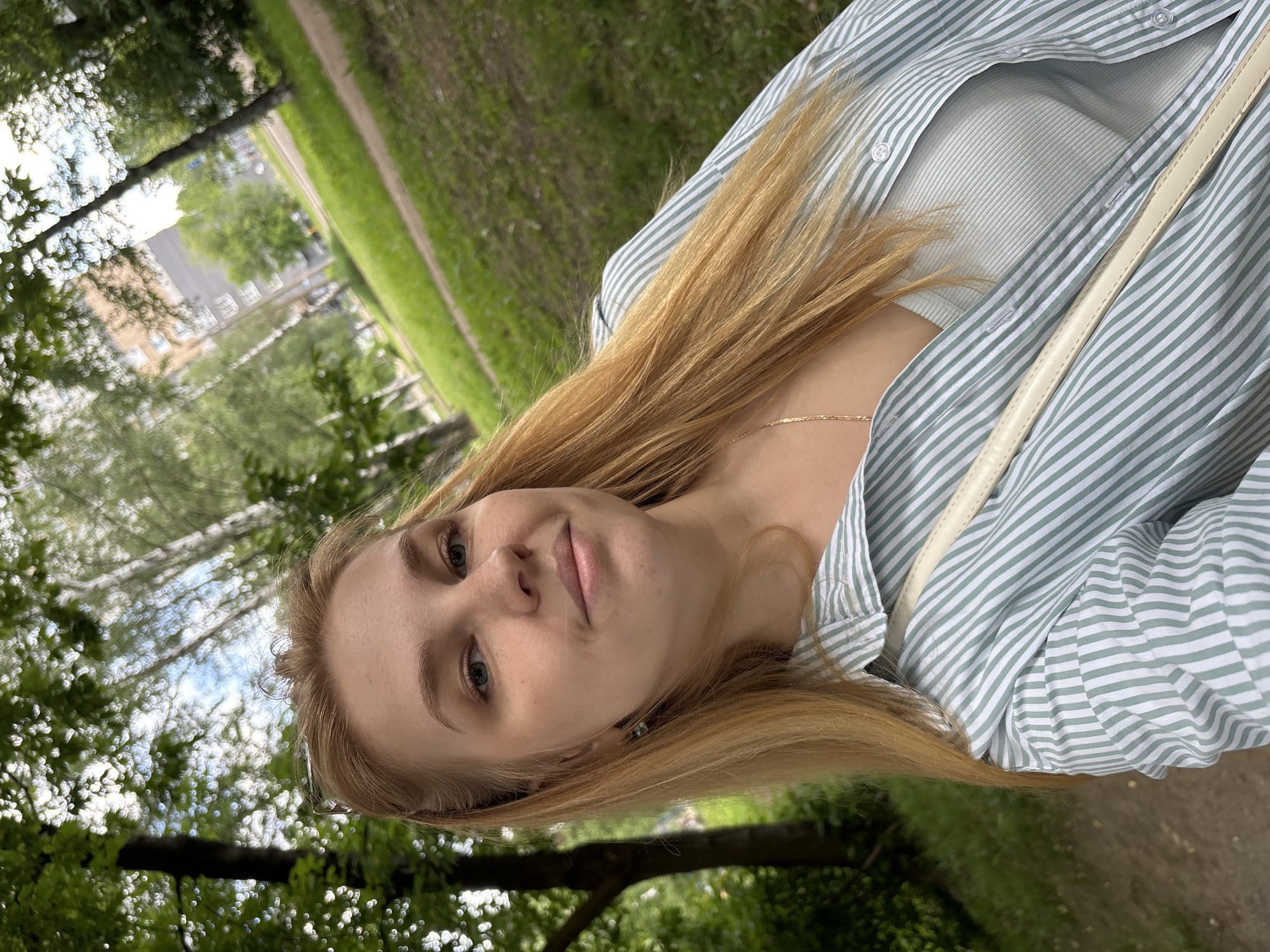 A young woman is featured in a rotated portrait-style photograph, set outdoors in a park or green area, likely in Rusanovo, Russia. She has long, straight, light brown to blonde hair that cascades over her shoulders. Her skin is fair, and she has light-colored eyes, looking directly at the camera with a composed, neutral expression.
She is dressed in a light green and white striped button-down shirt, which is partially unbuttoned, revealing a light-colored (possibly white or very light green) ribbed tank top or camisole underneath. A delicate gold-colored chain necklace is visible around her neck. A light-colored strap, likely from a handbag or shoulder bag, crosses her right shoulder.
The background is rich with green foliage and trees, some appearing as bokeh due to a shallow depth of field. A narrow, unpaved path or dirt road is visible on the right side of the background. In the further distance, through the trees, blurred outlines of buildings or structures suggest an urban or suburban environment adjacent to the green space. The lighting indicates a bright daytime setting. There is no visible text in the image.