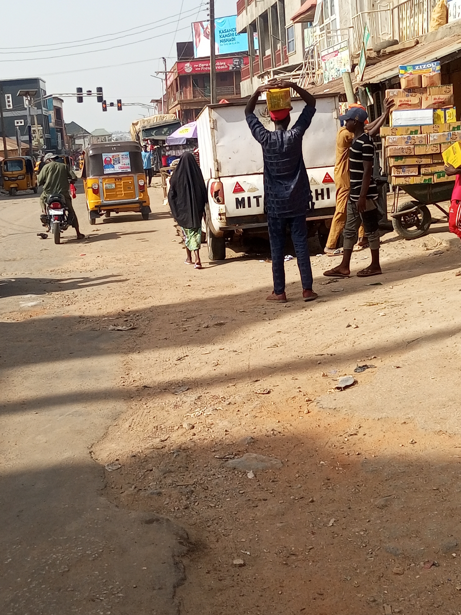 Here is a forensic description of the image:
Location: City of Maiburiji, Nigeria.
Scene: A dusty street scene on a sunny day.
Subjects:
* Several people are present. One man is carrying a yellow container on his head, wearing a dark blue shirt and jeans. Another man is wearing a striped shirt and a cap.
* A woman in a dark hijab is walking near a white truck. A person in a yellow robe is visible partially behind the truck.
* A motorcyclist is riding on the left side of the road.
Objects:
* Several yellow "kekes" (auto rickshaws) are driving on the road.
* A white truck with the partial word "MITSUBISHI" on its rear.
* A hand-pulled cart loaded with boxes.
* Traffic lights are visible in the background.
* Advertising billboards are on buildings in the background.
Environment:
* The street is dusty and uneven.
* Buildings with shops are visible along the street.
* The sky is clear.