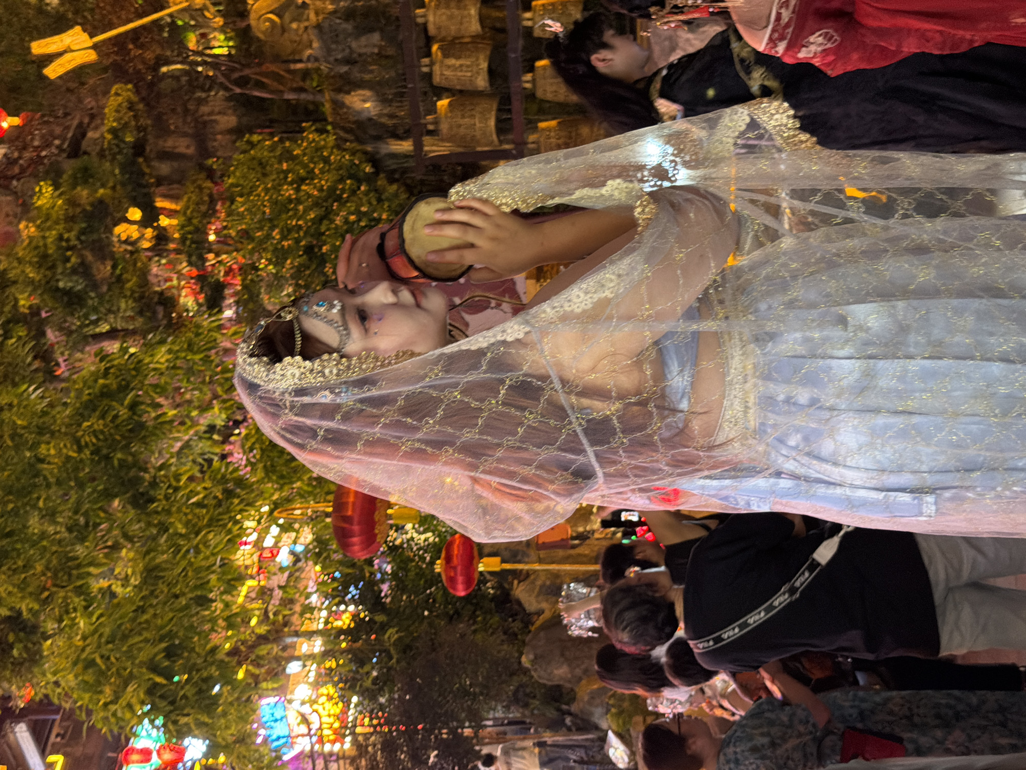 The image shows a group of people outdoors at night, likely at a festival or cultural event in Yuzhong District, China. The primary subject is a person in the foreground dressed in elaborate attire, possibly a costume or traditional garment, with a sheer veil adorned with gold patterns covering their head and shoulders. They are wearing a decorative headpiece with blue gems.
Behind this individual, other people are visible, some appearing to be watching or participating in an event. There are red lanterns hanging in the background, contributing to a festive atmosphere. The setting seems to be an outdoor area with trees and decorative lights. The lighting suggests it is nighttime. One person in the background is wearing a black shirt with white text that appears to be "FILA" repeated. The overall mood seems festive and celebratory.